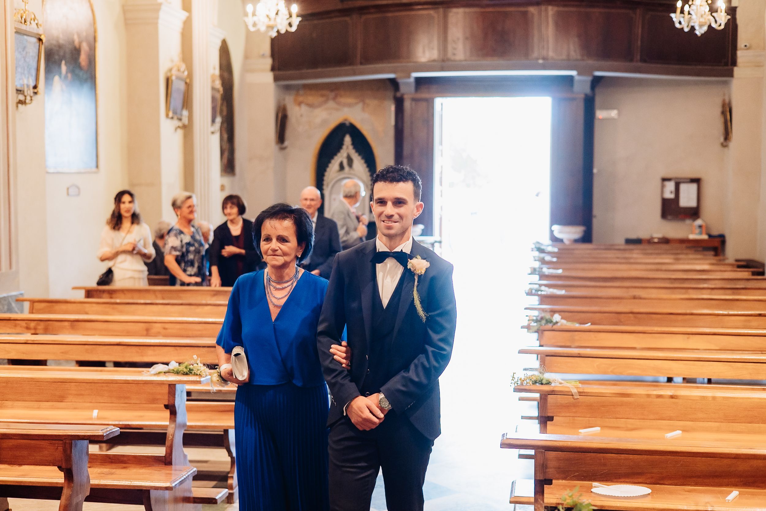 Wedding party in navy blue attire poses inside a church with wooden pews and crystal chandeliers.