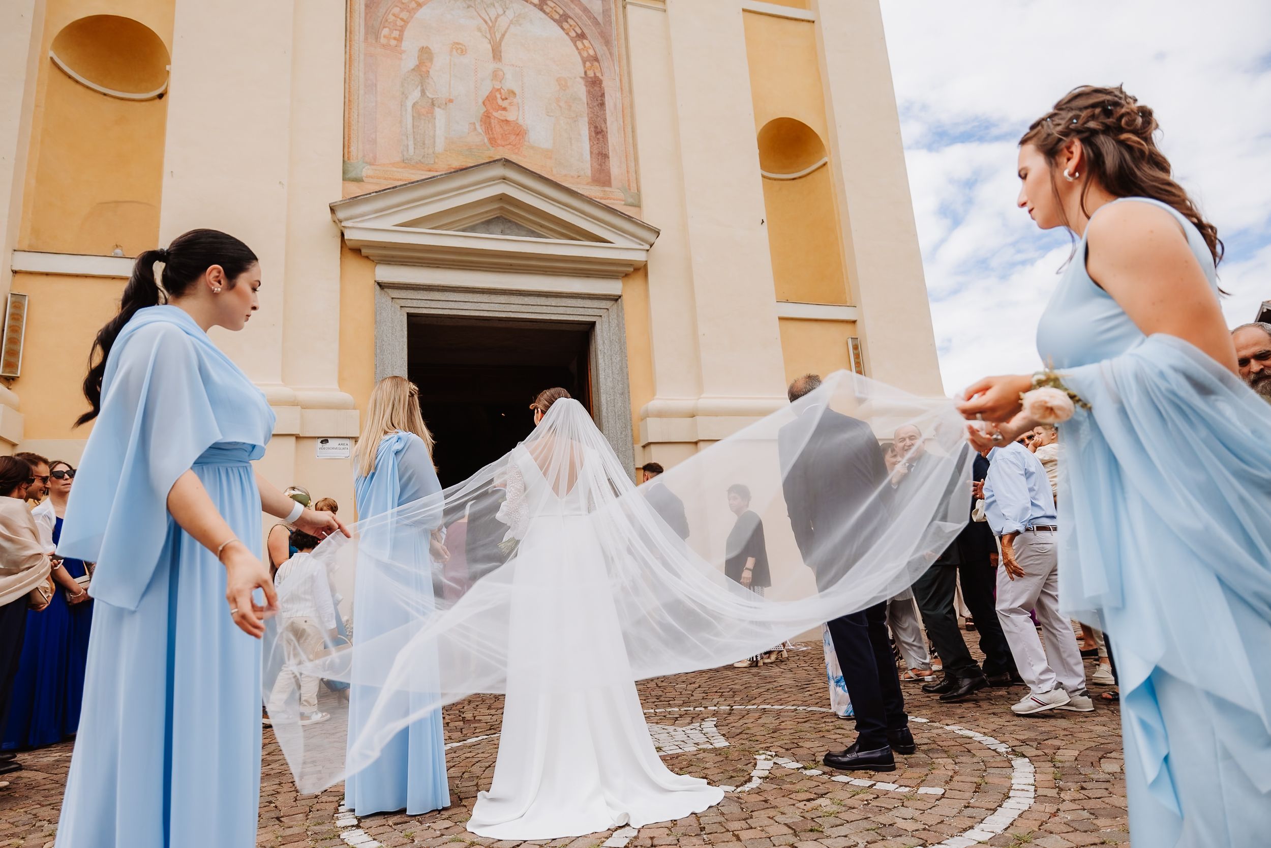 Wedding party in light blue dresses helps adjust bride's flowing veil outside a church.