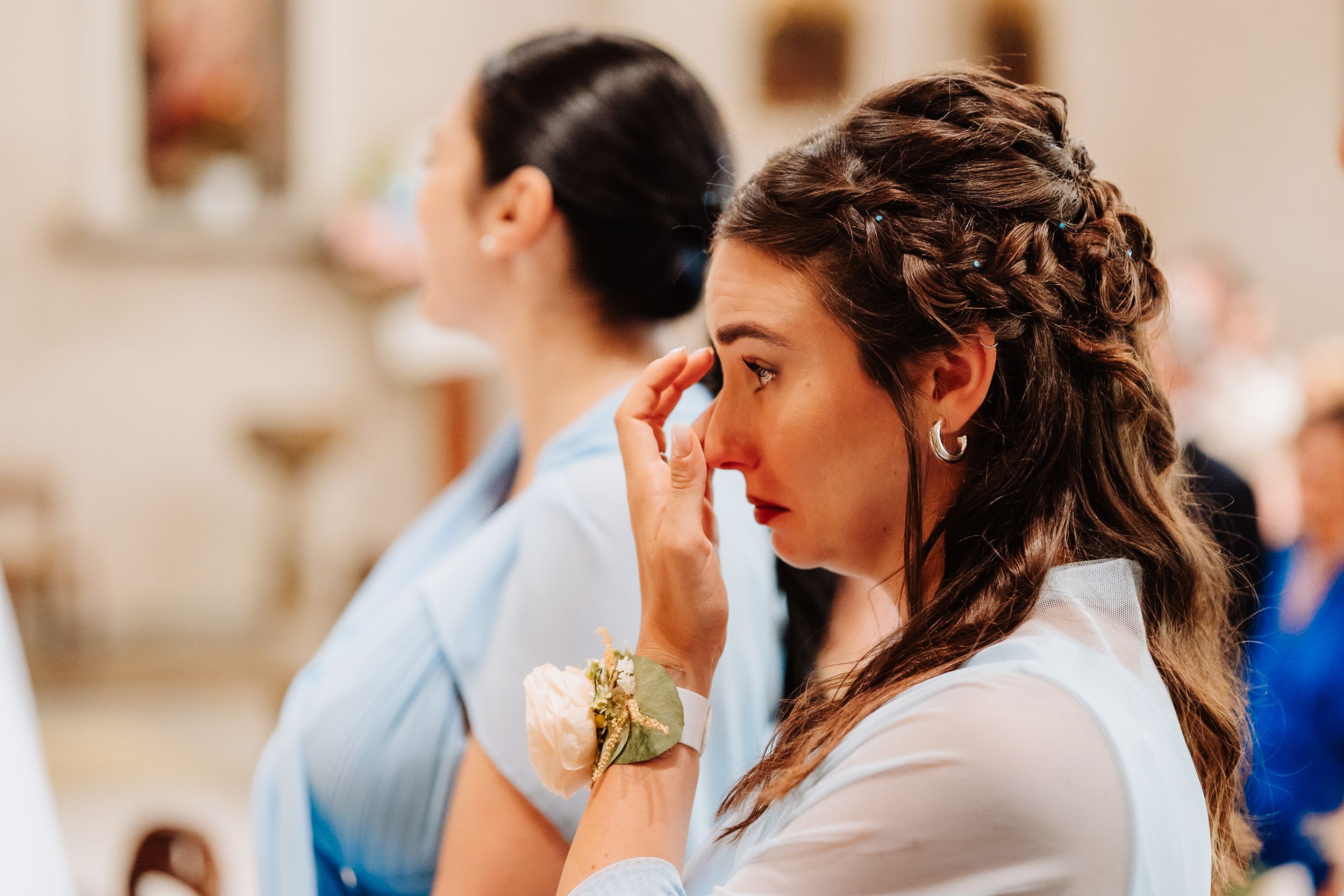 An emotional guest wipes away tears during a wedding ceremony, wearing a white blouse with an elegant braided updo hairstyle.