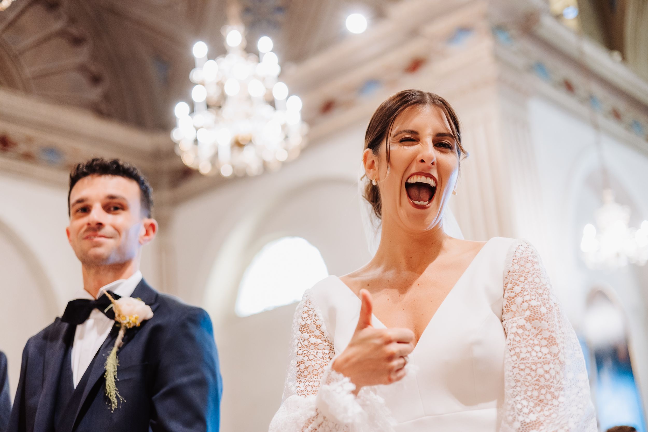 Wedding guests laugh joyfully during a reception celebration in an elegant ballroom with crystal chandeliers.