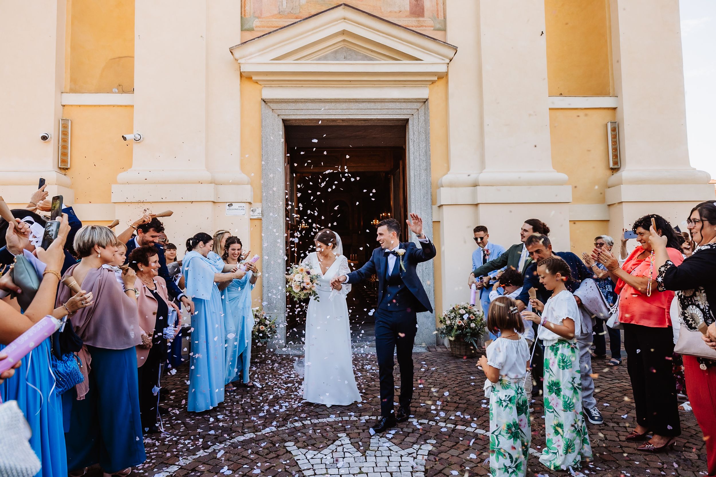 Wedding party celebrates with confetti outside a yellow church building on a sunny day.