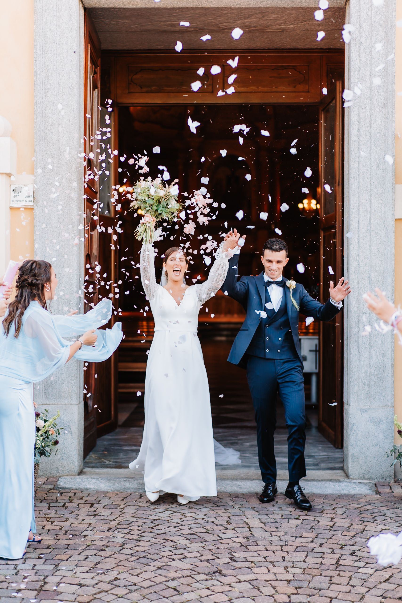 Wedding party celebrates with confetti being thrown outside a rustic stone building entrance.