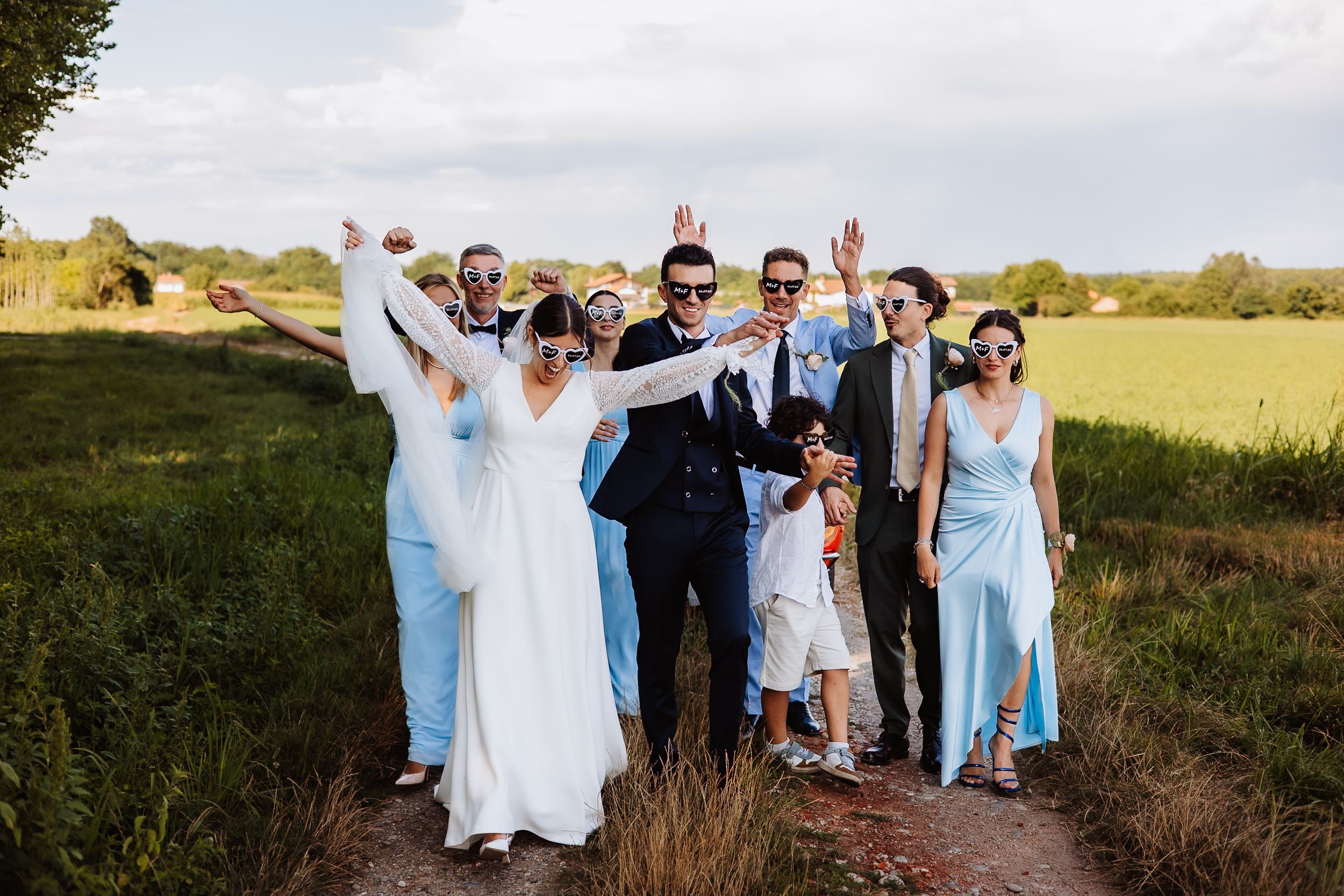 Wedding group poses together in a sunny field with light blue and white attire.