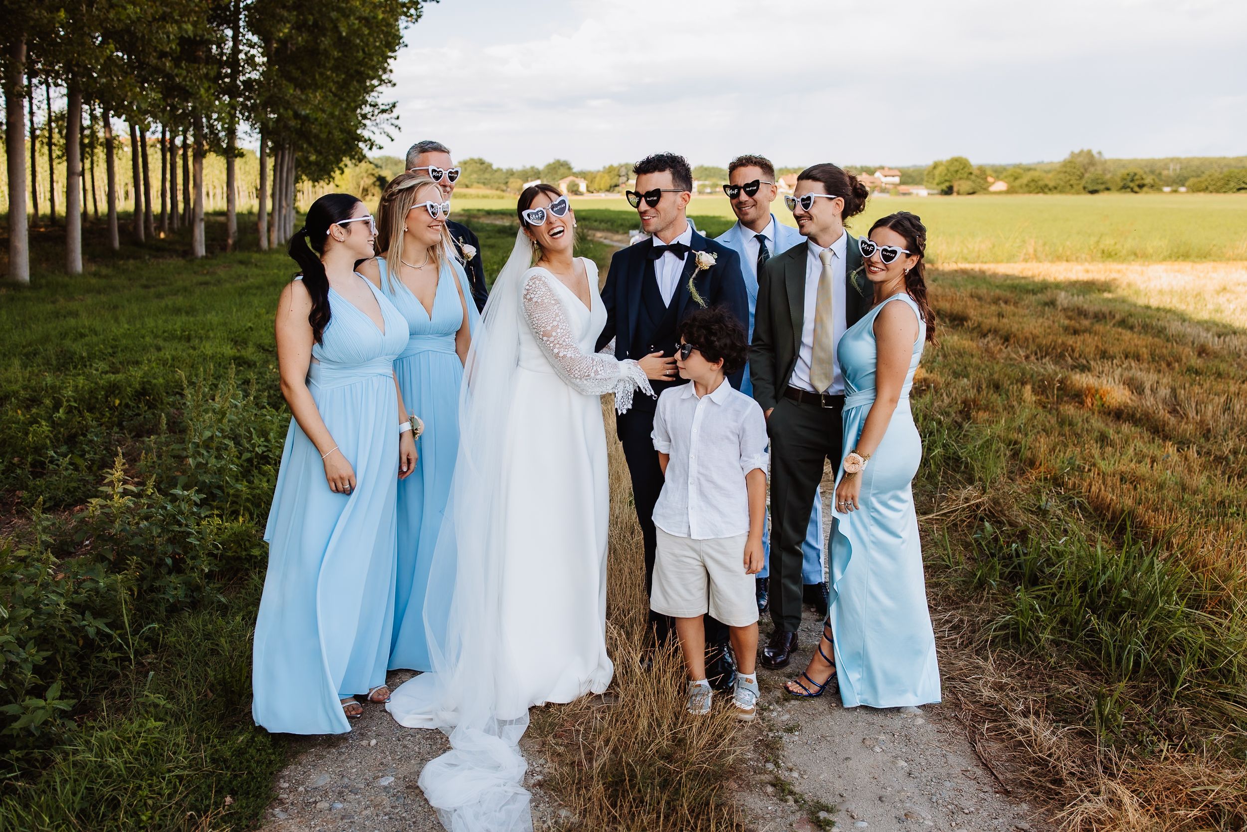 Wedding party in light blue bridesmaid dresses and formal attire pose together on a rustic outdoor path at sunset.
