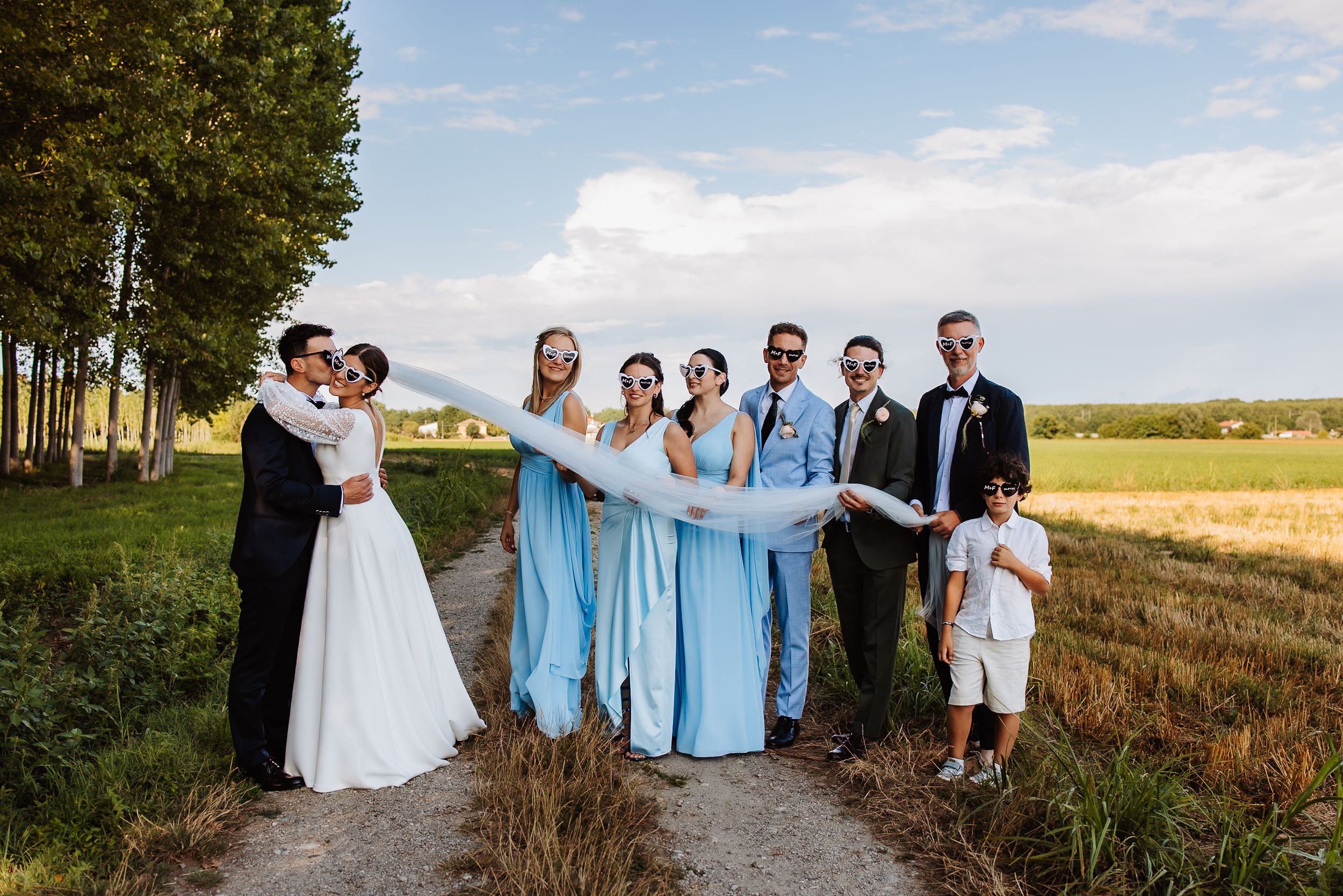 Wedding party in formal attire poses together on a dirt path with golden fields and trees in the background.