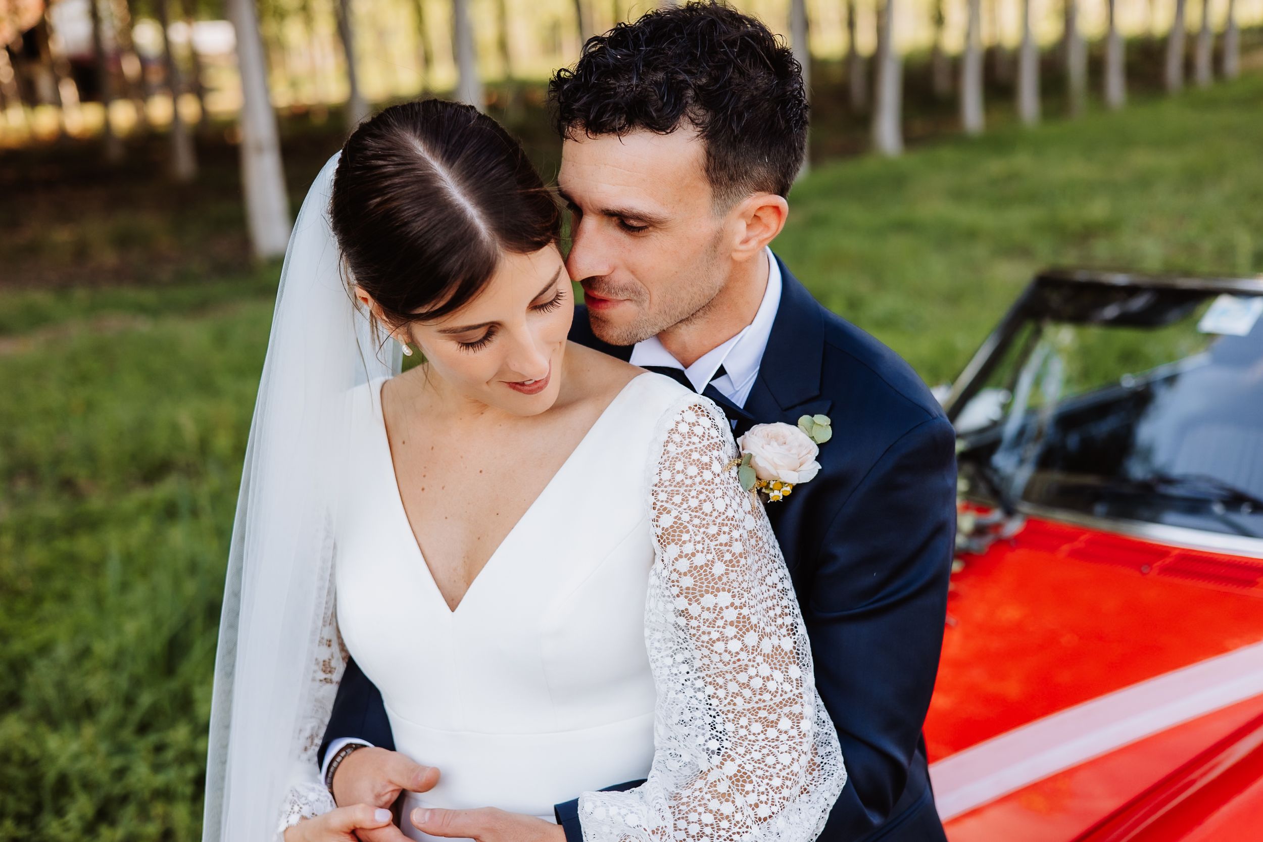 Couple in wedding attire embraces near a red vintage car in an outdoor setting.