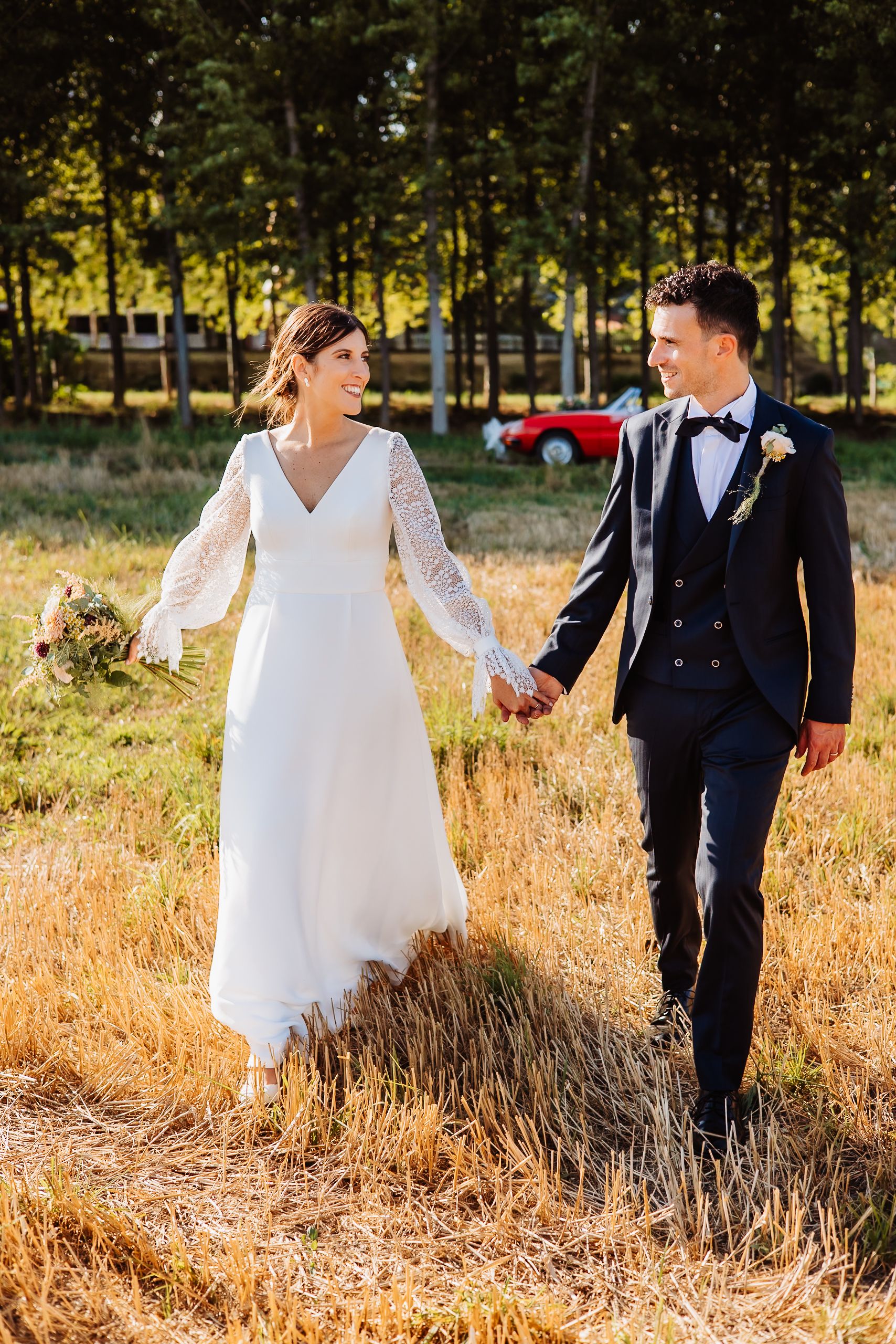 A couple in wedding attire walks hand in hand through a golden field at sunset with trees in the background.