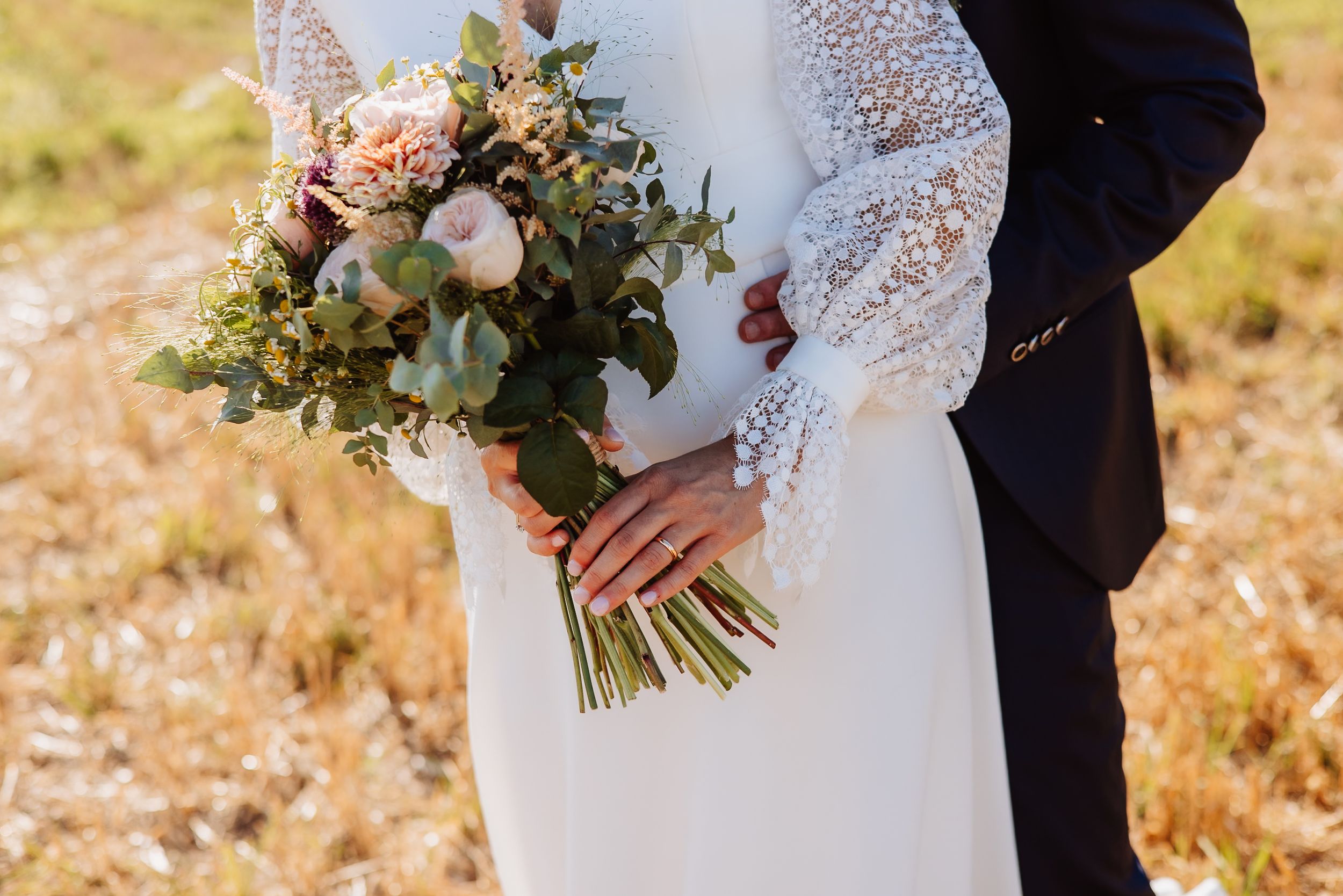 A close-up detail shot of a bride and groom's hands with the wedding bouquet in a rustic outdoor setting.