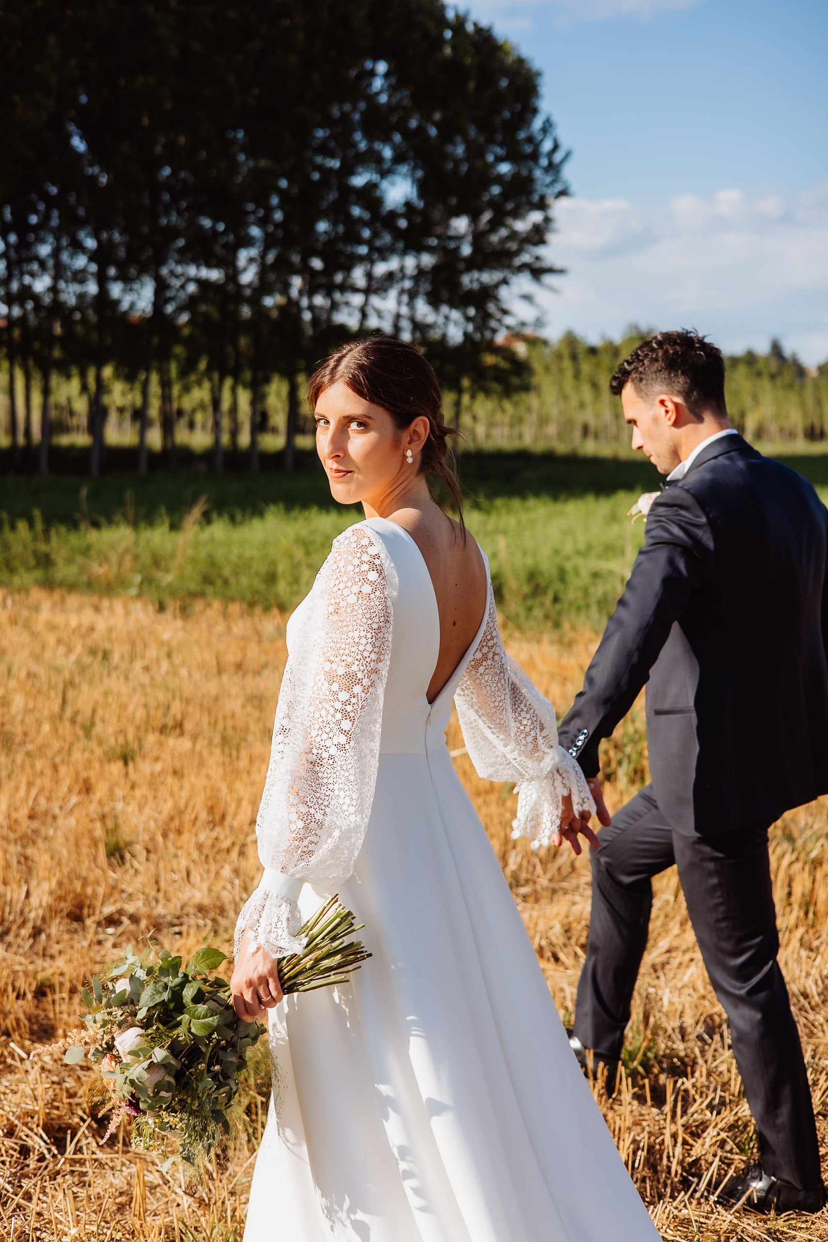 Newlywed couple walks through golden wheat field during sunset wedding photoshoot.