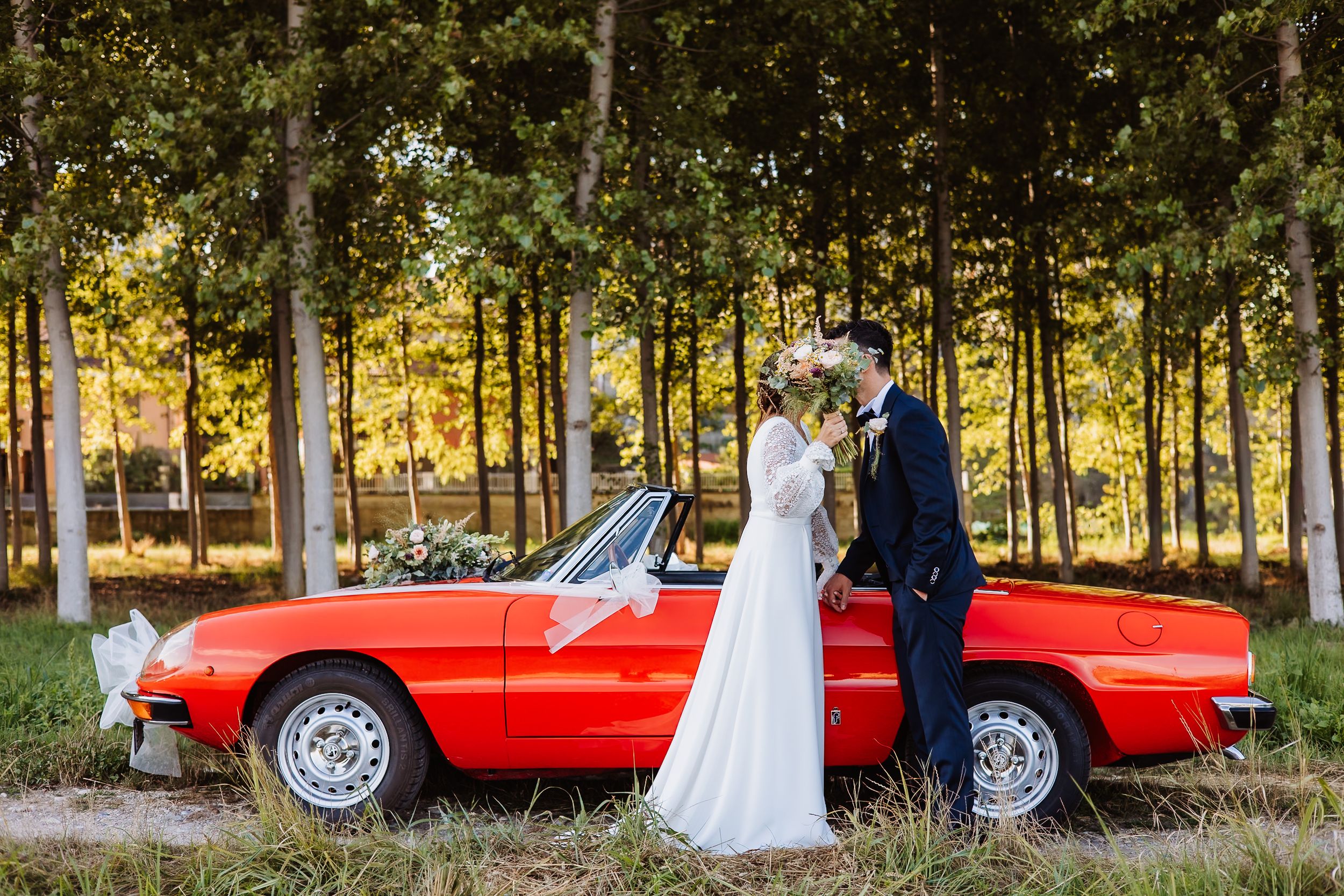A red vintage convertible car parked in a wooded area serves as a backdrop for a wedding photo session.