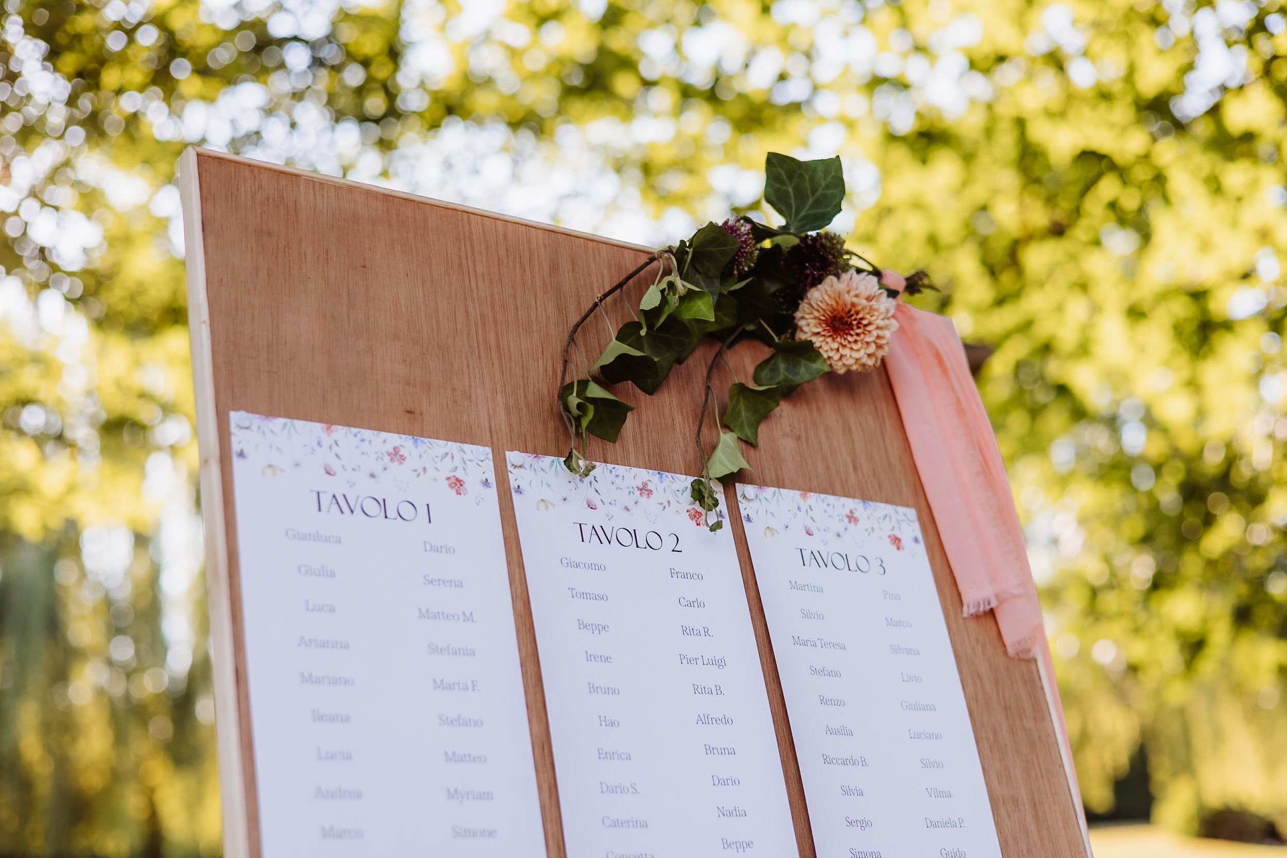 Wedding seating chart display board decorated with floral accents against a sunlit natural backdrop.