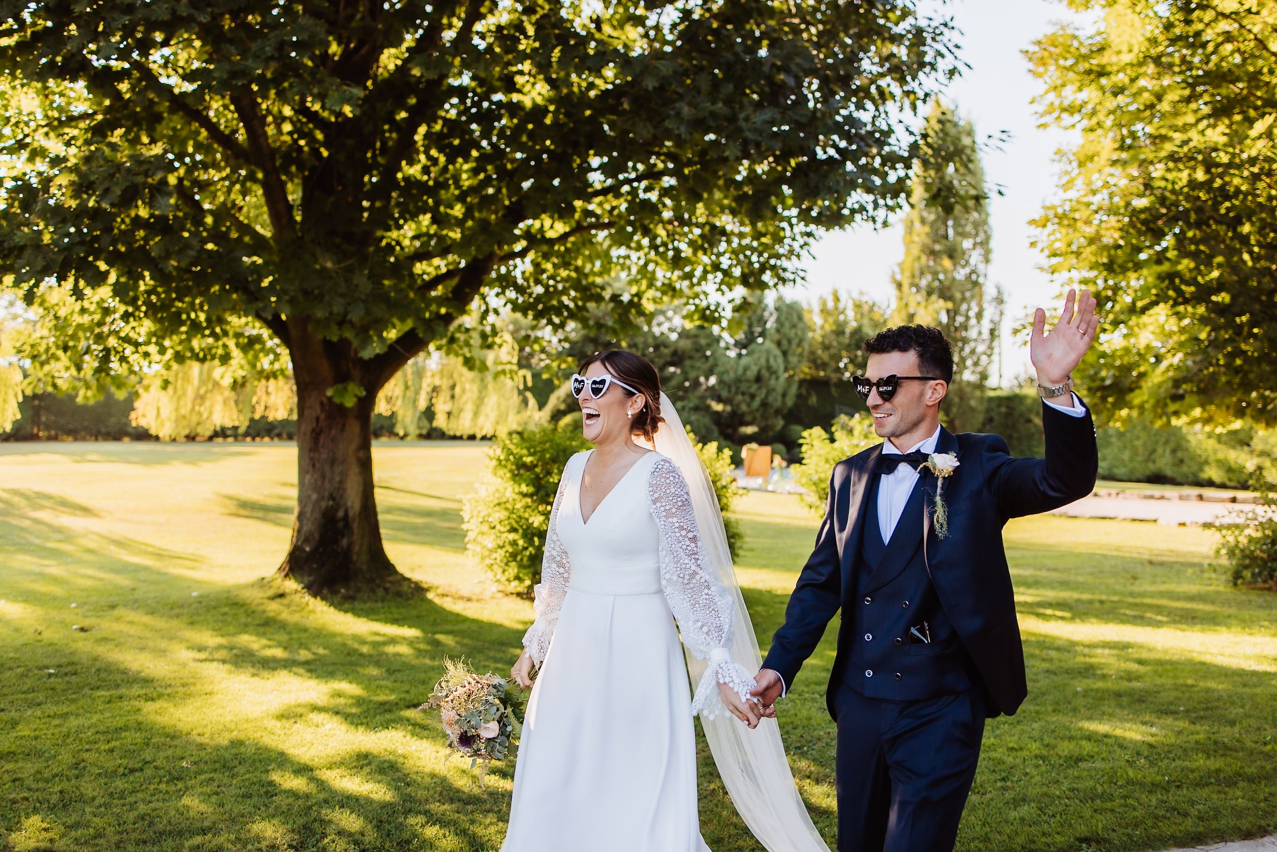A couple in wedding attire walks together on a sunny autumn day in a park setting with golden leaves.