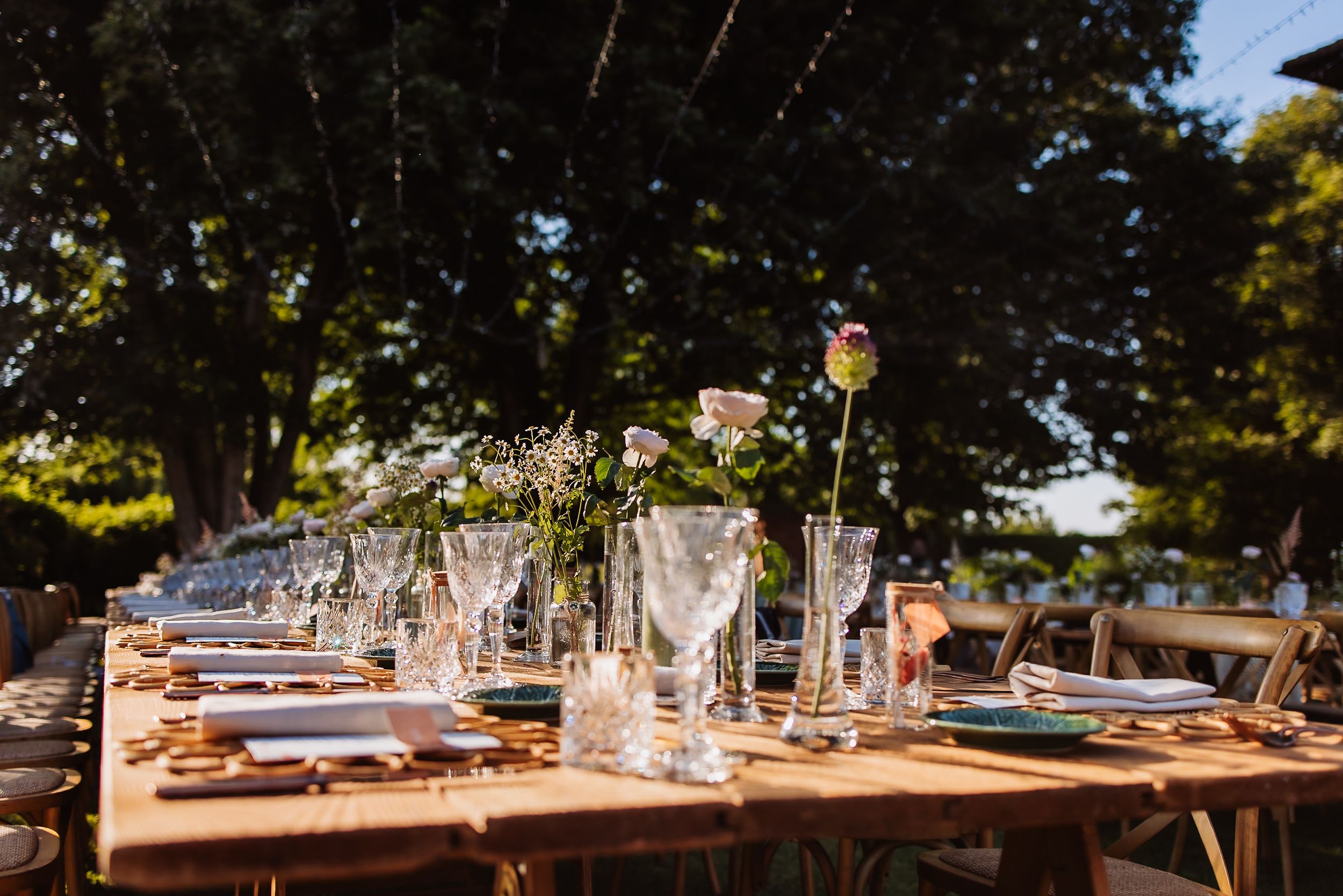 Long wooden tables with place settings and floral arrangements are set up outdoors under tall trees for a wedding reception.