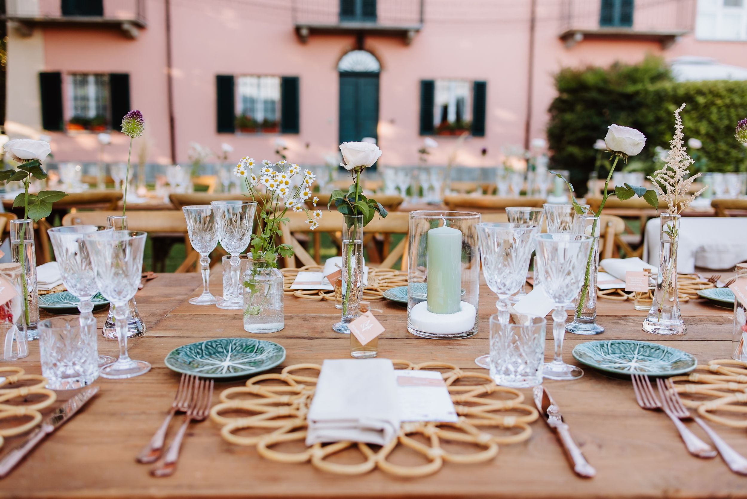 Elegant outdoor table setting with rattan placemats, crystal glassware, and delicate floral centerpieces against a pink building.