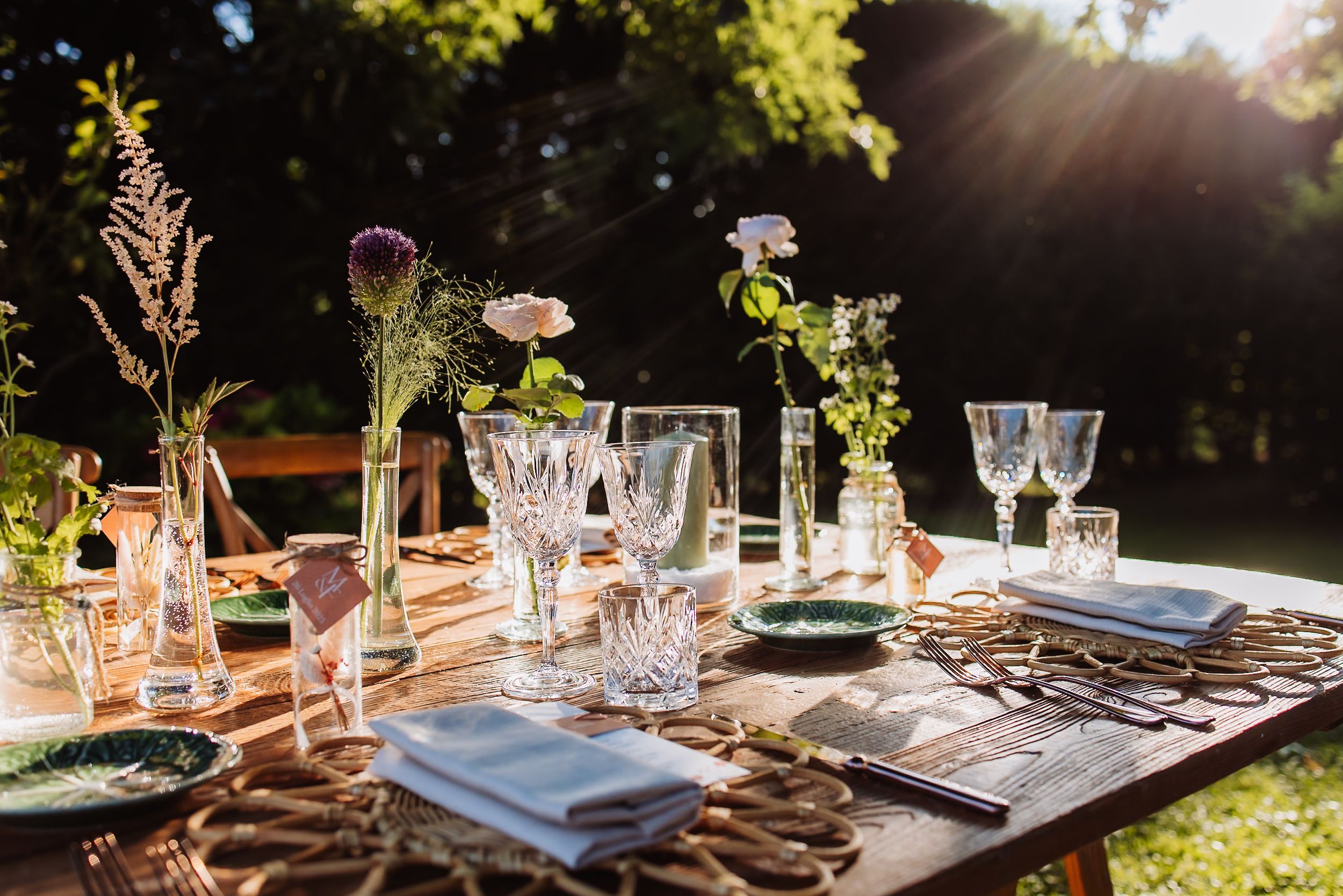 Elegant place settings with crystal glassware and floral centerpieces on wooden tables at sunset outdoor reception.