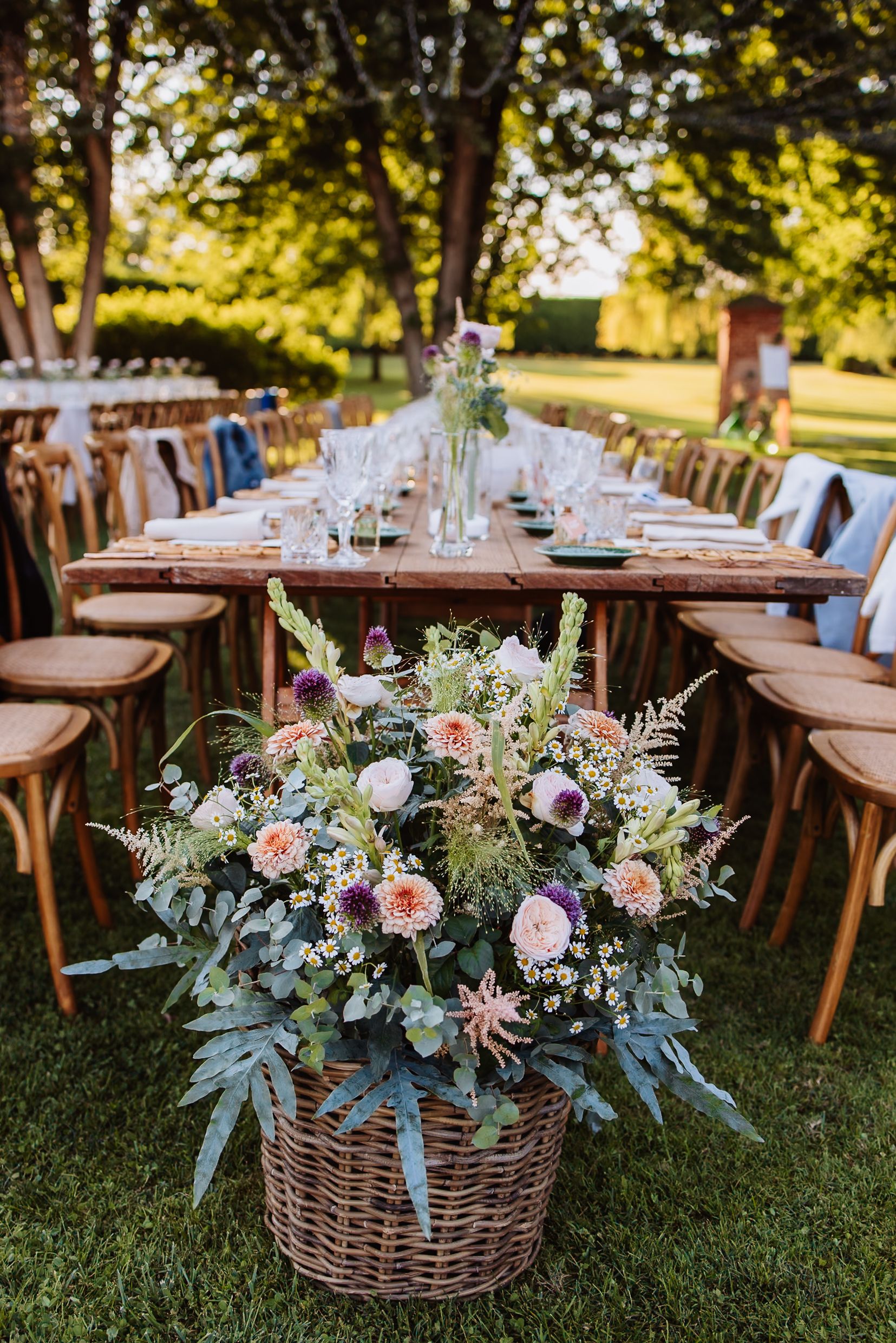Elegant outdoor wedding reception tables decorated with floral centerpieces in wicker baskets under sunlit trees.