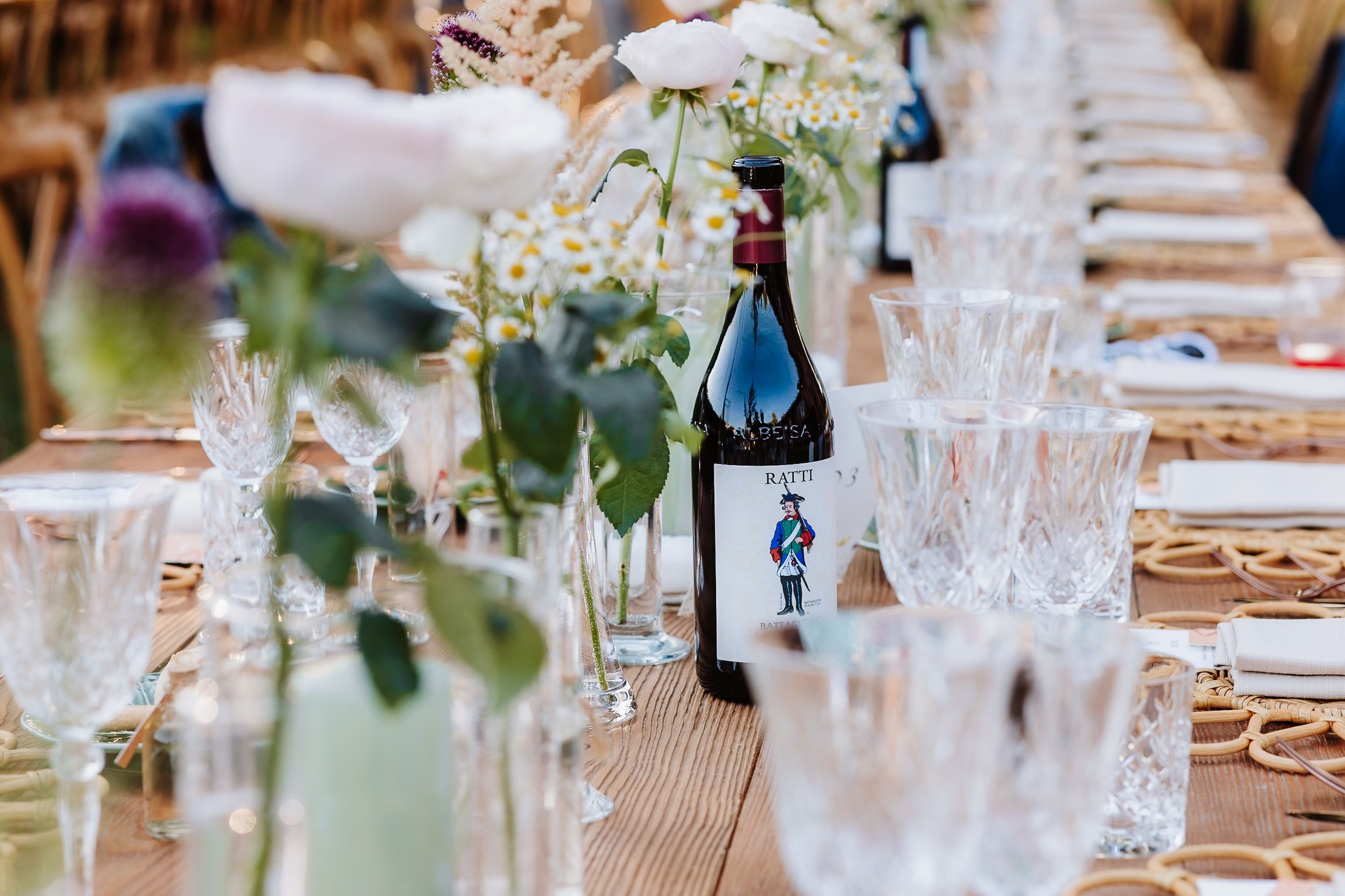 Wedding reception table with crystal glassware, wine bottles, and delicate floral arrangements on wooden surface.