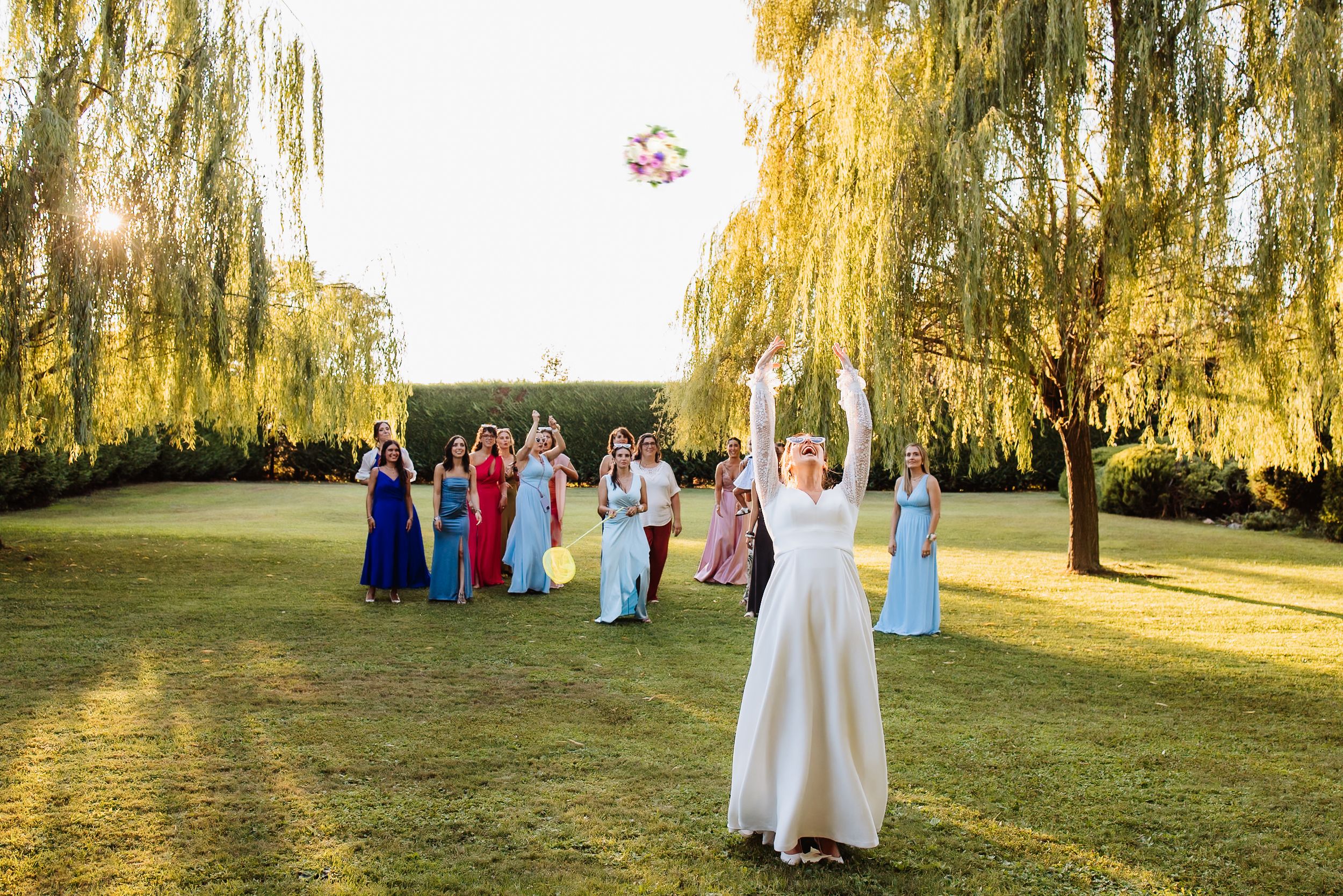 Wedding celebration group poses beneath weeping willow trees during golden hour sunset.