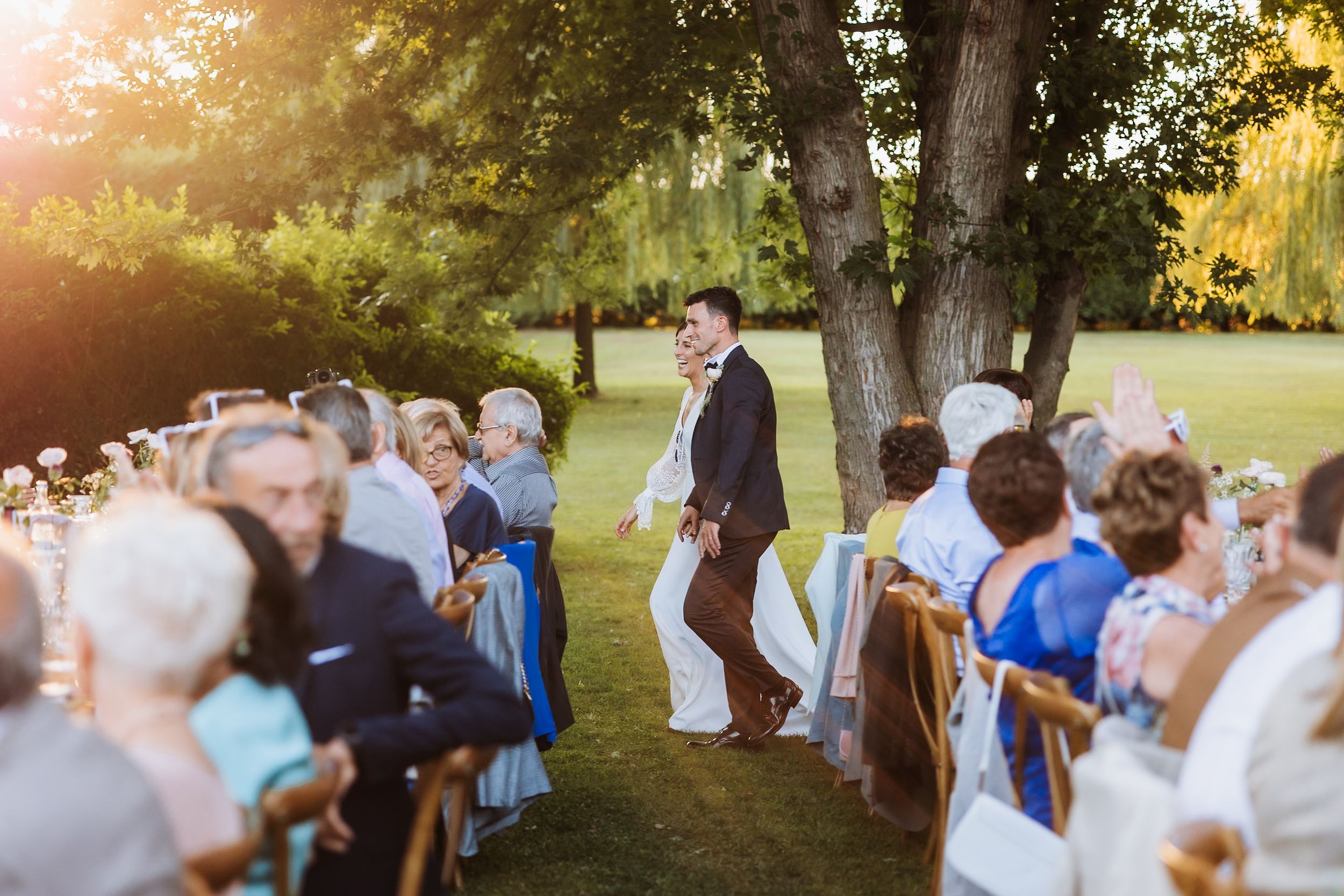 Outdoor wedding ceremony in a sunlit garden with guests seated on white chairs.