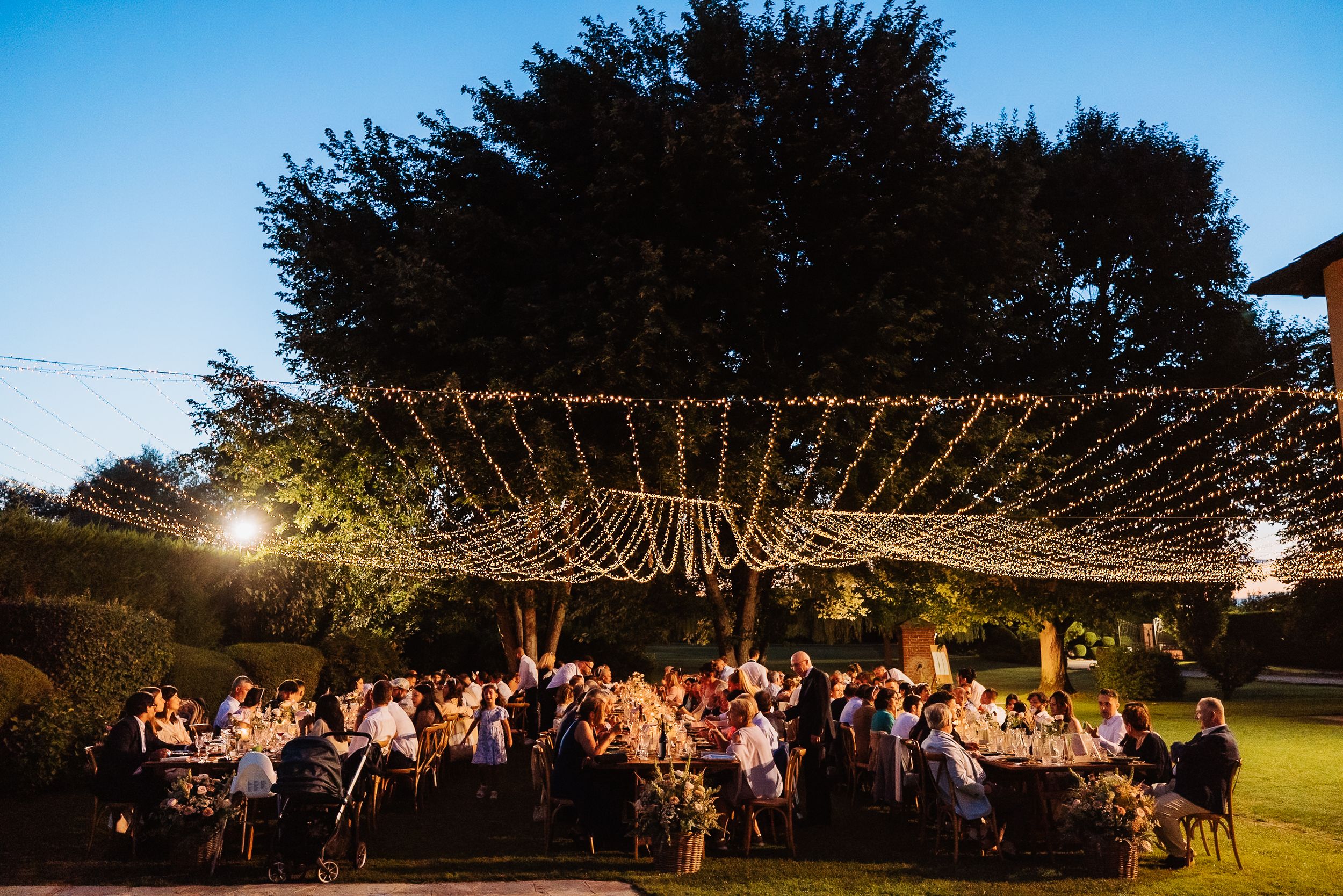 String lights illuminate an outdoor evening wedding reception with guests dining under a canopy.