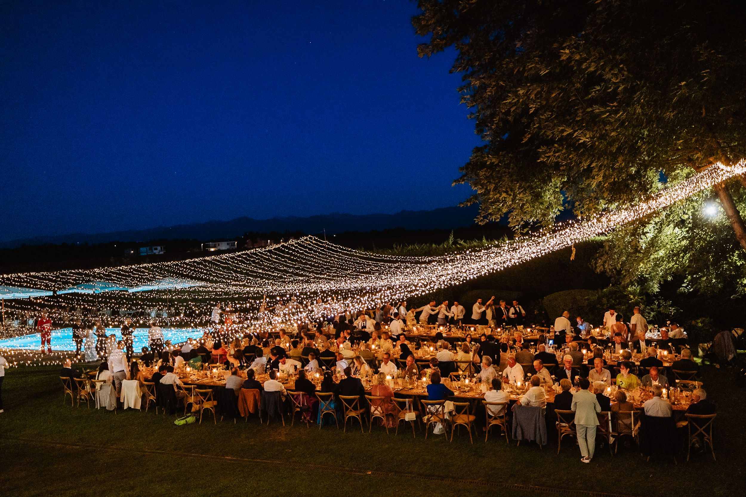Panoramic view of a large outdoor wedding reception at night with string lights illuminating the crowd.