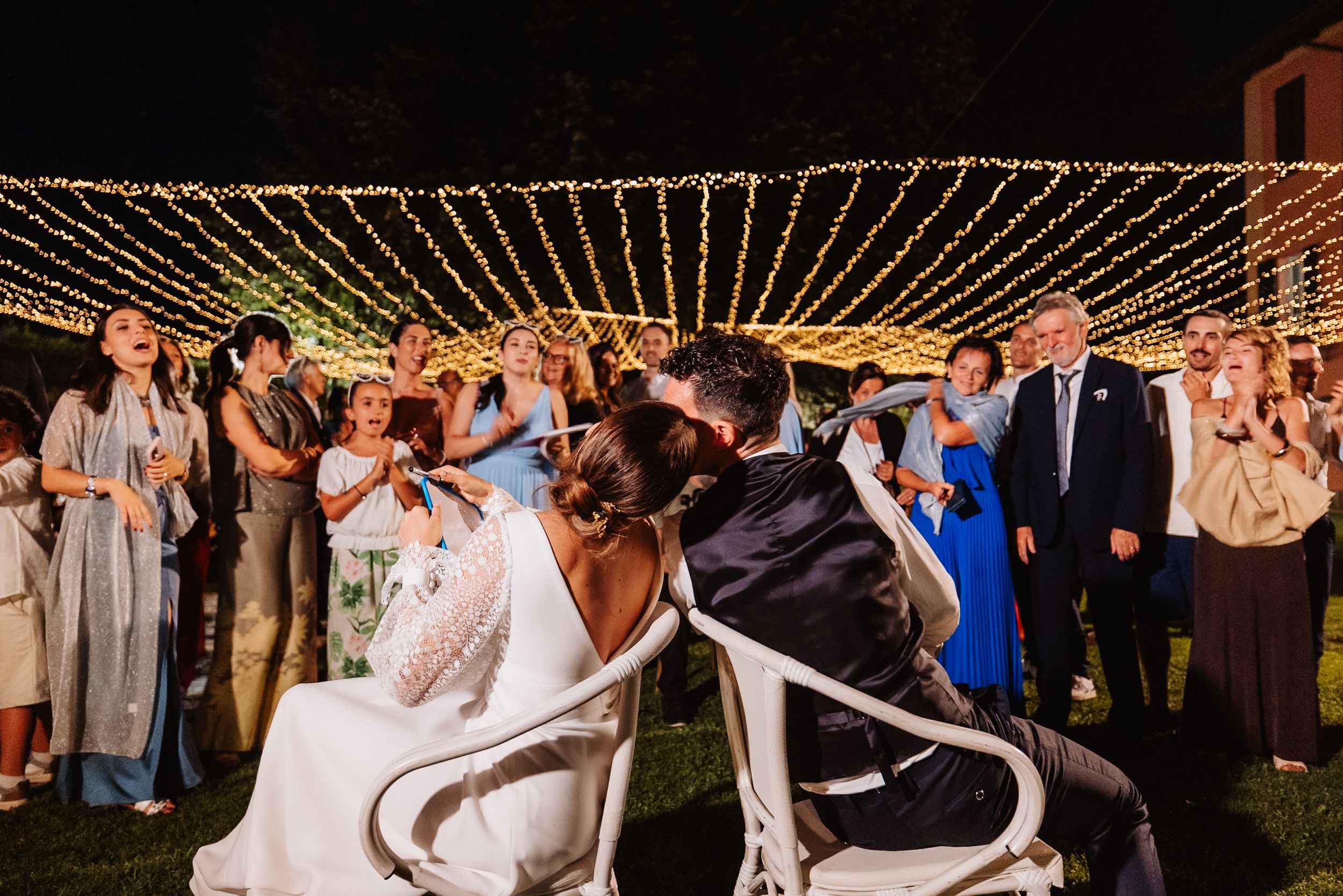 Wedding guests dance and celebrate under a canopy of twinkling string lights at an outdoor evening reception.