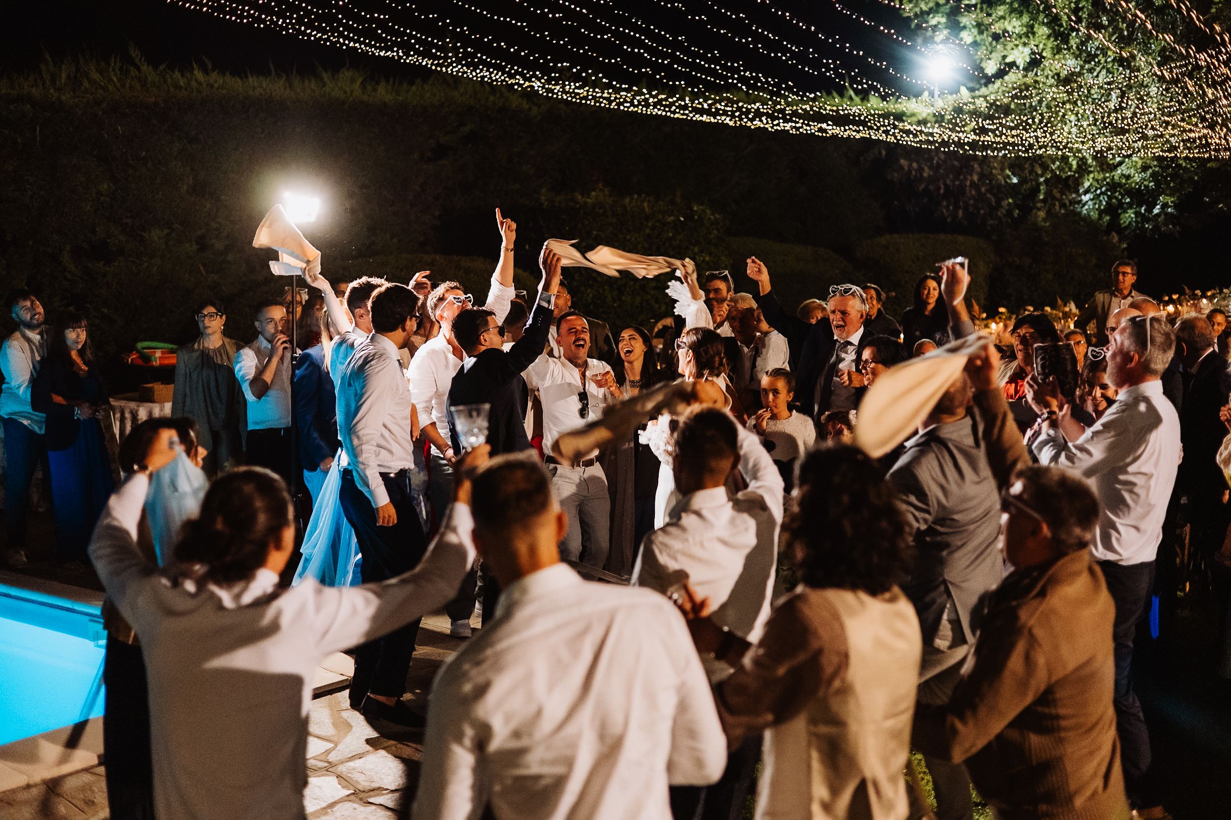 A lively crowd dances under illuminated string lights at a nighttime outdoor celebration.