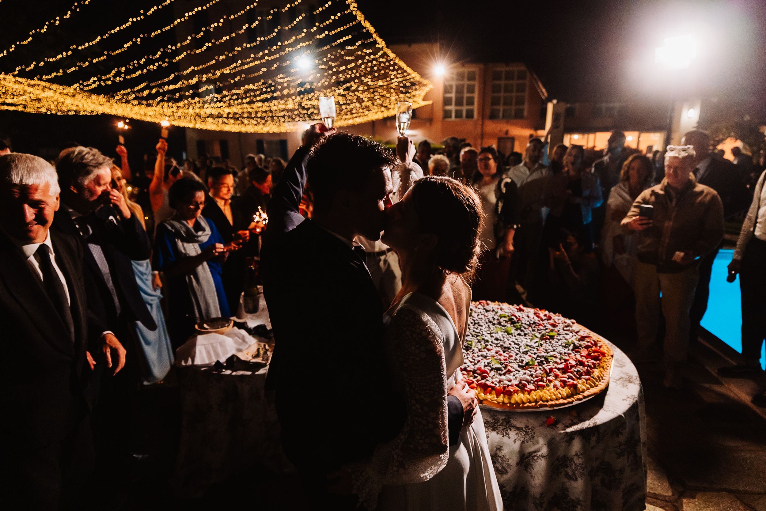 Nighttime wedding reception with string lights overhead and crowd gathered around illuminated dessert table.