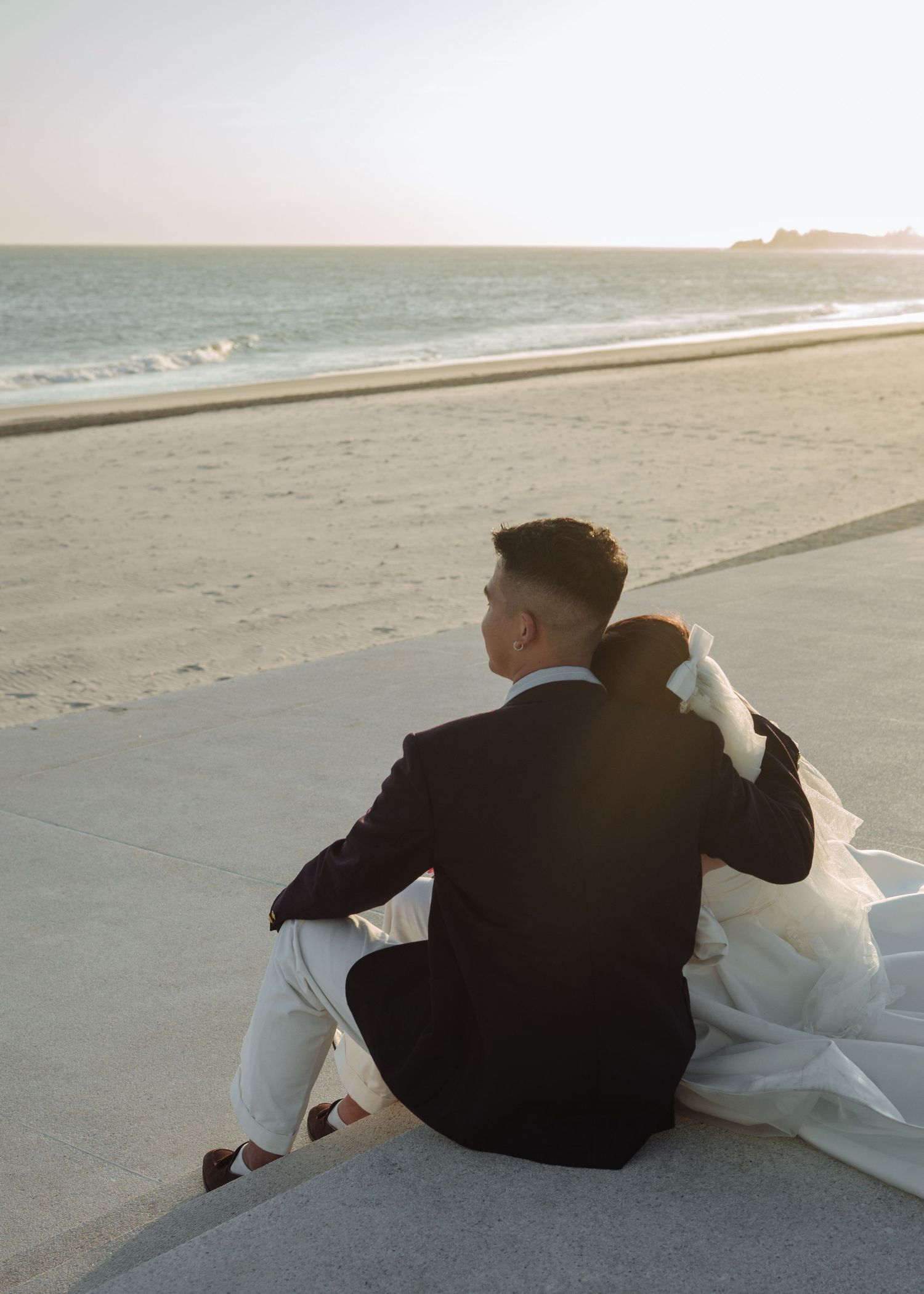 Bride and groom sitting together on beachside promenade watching golden sunset over sandy shore.
