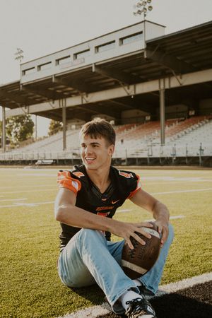 Football player in black uniform sitting casually on field with stadium in background.