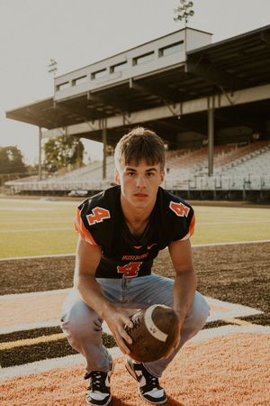 Football player in black jersey crouching on field near stadium bleachers at sunset.