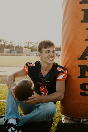 Football player in black jersey posing with ball on sunny field.