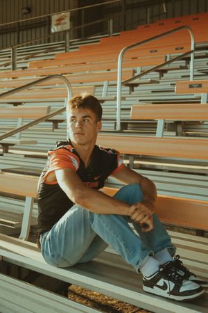 Football player in casual attire relaxing on bleacher seats in an indoor stadium.