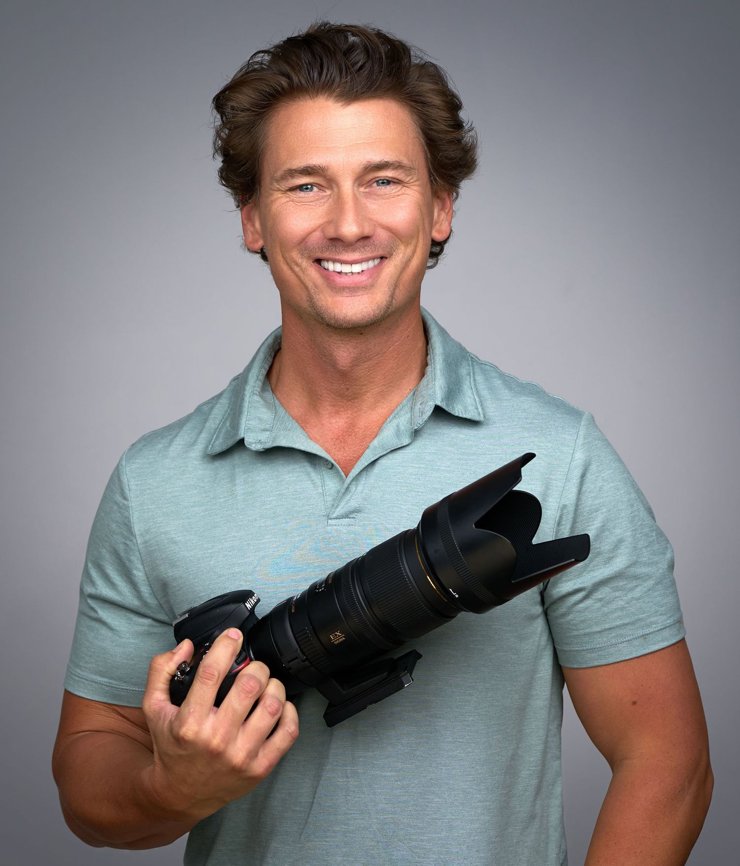 Smiling man in teal polo shirt holding a professional DSLR camera with telephoto lens against gray background.