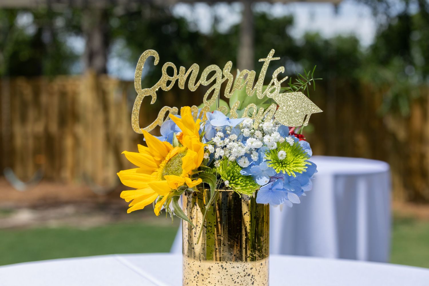 A floral centerpiece with yellow sunflowers and blue daisies in a gold glitter vase with a 'Congrats' sign on top.