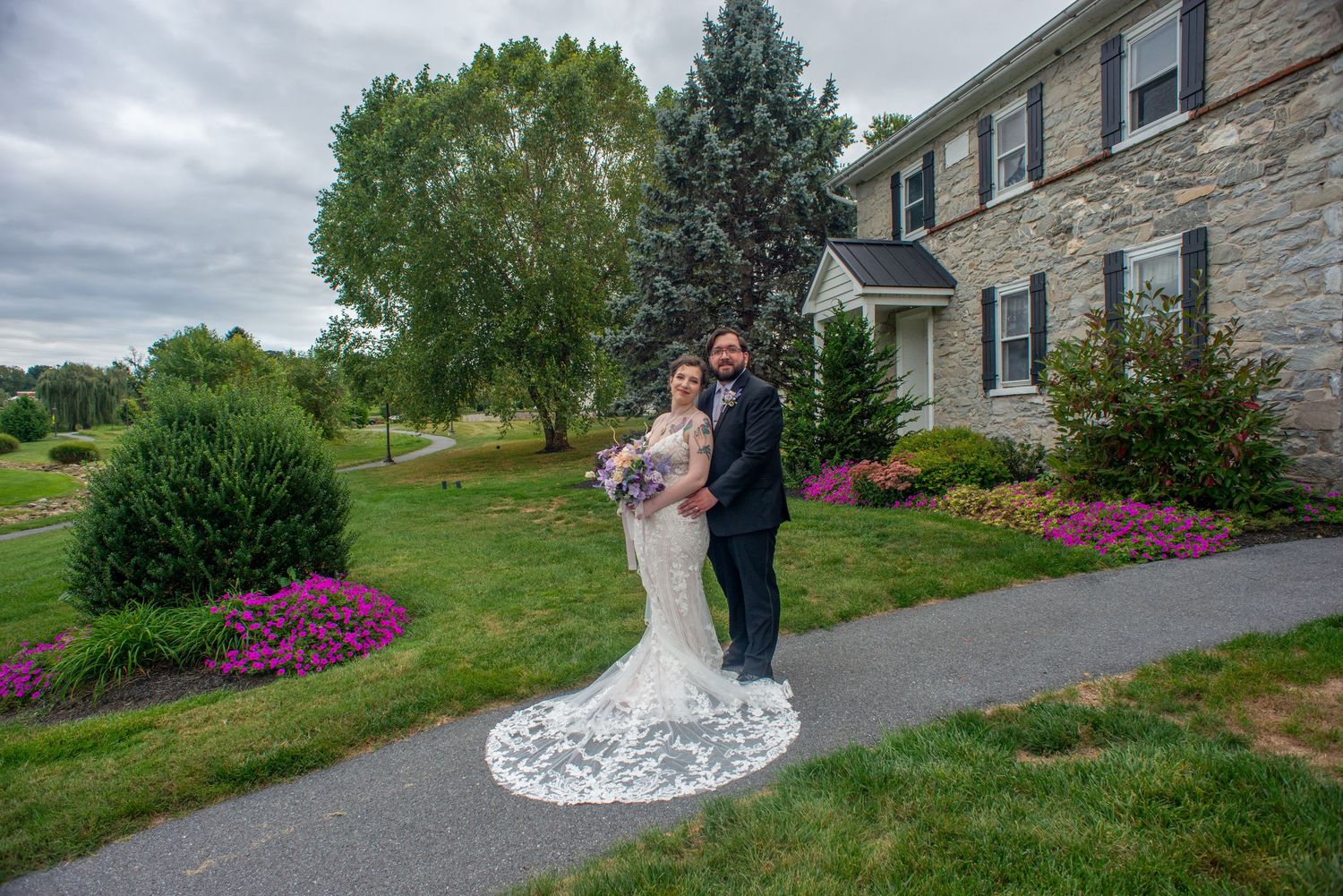 A couple poses for outdoor wedding photos in front of a stone house with blooming azalea bushes and green landscaping.