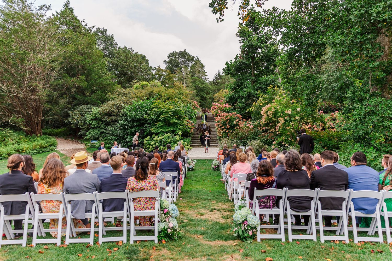 Panoramic view of an outdoor wedding ceremony setup with white chairs arranged in rows surrounded by lush greenery.