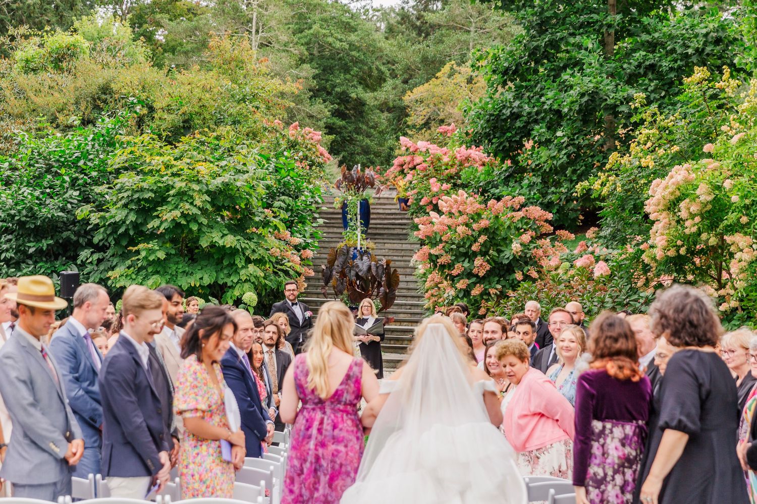 Wedding guests gather outdoors in a garden setting during a sunny celebration.