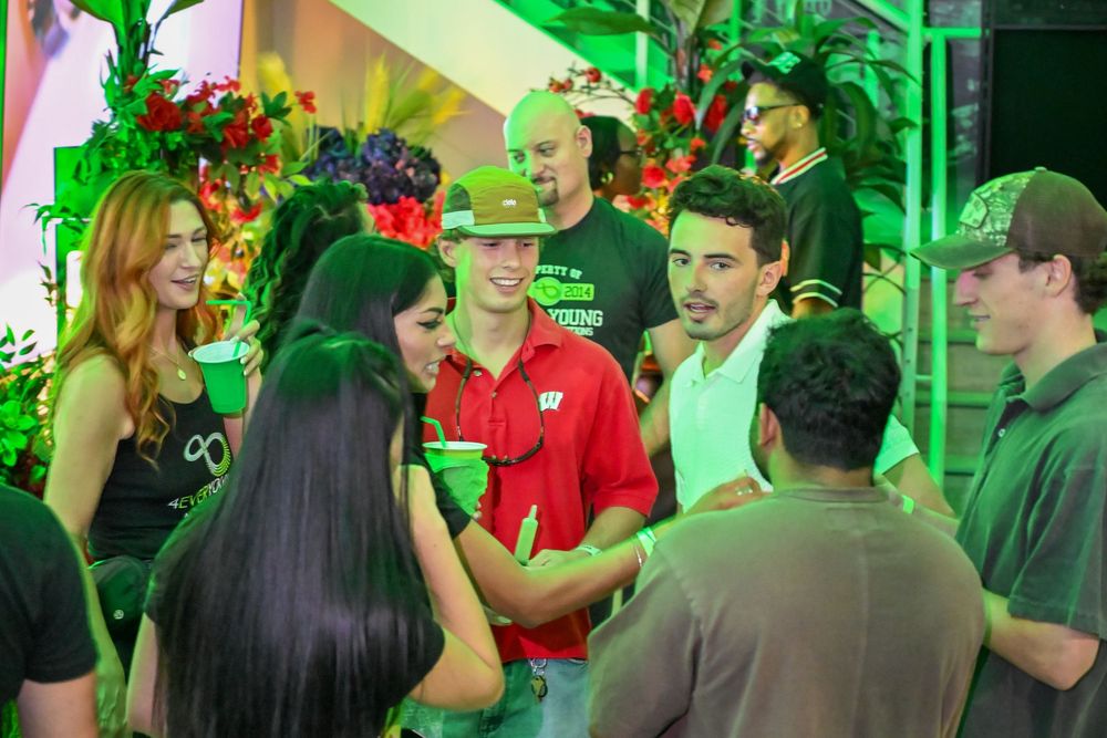 People gather at a bar or nightclub illuminated by bright green lighting while socializing and drinking.