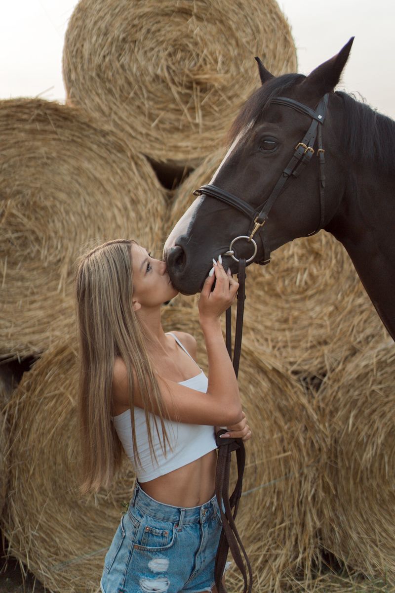A person in denim shorts shares an affectionate moment with a dark horse against a backdrop of hay bales.