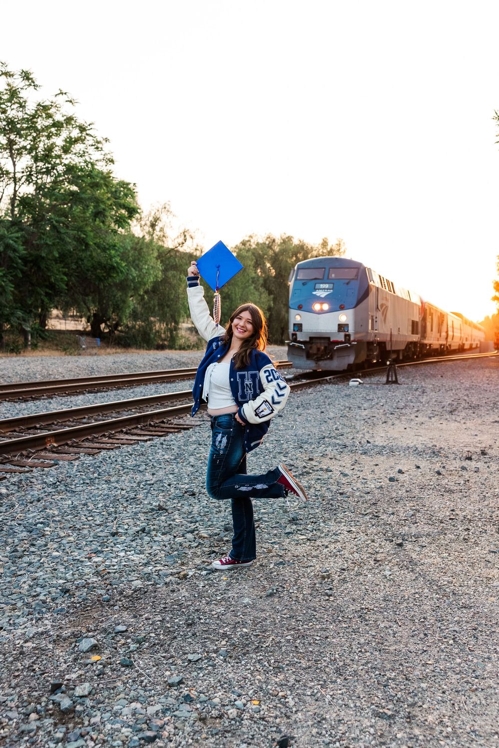 A graduate poses playfully near train tracks at sunset with a parked train in the background.