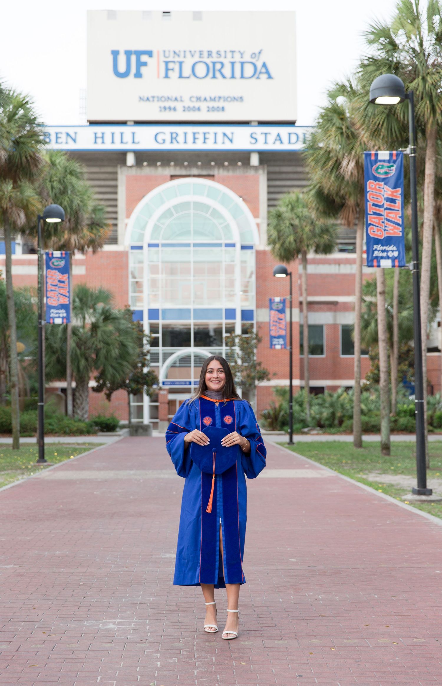 University of Florida graduate in royal blue cap and gown posing in front of Ben Hill Griffin Stadium.
