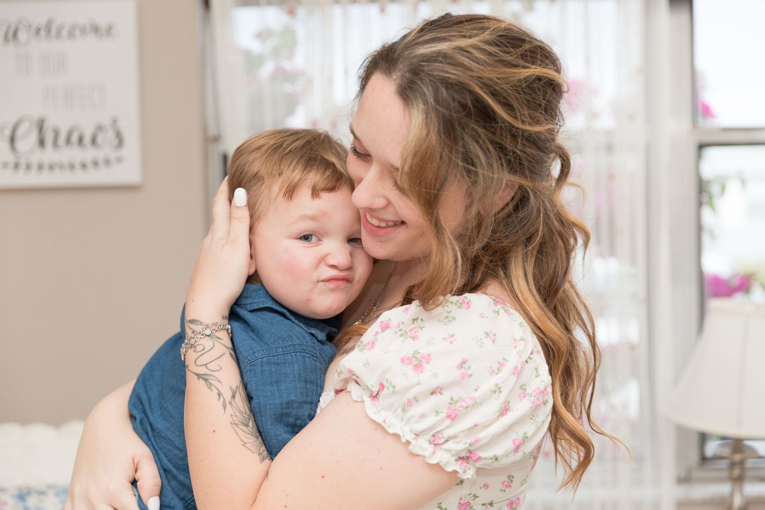 Mother with tattoos tenderly hugging her young toddler son indoors in a decorated room.