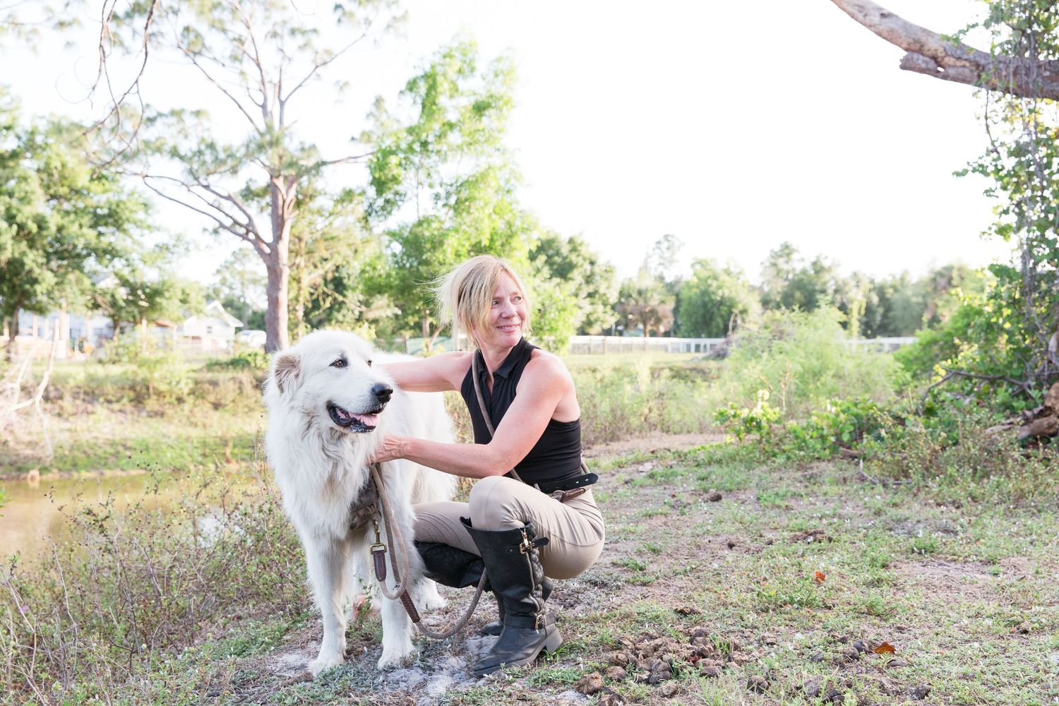 Blonde woman crouching beside a white dog outdoors near a pond in a natural setting.