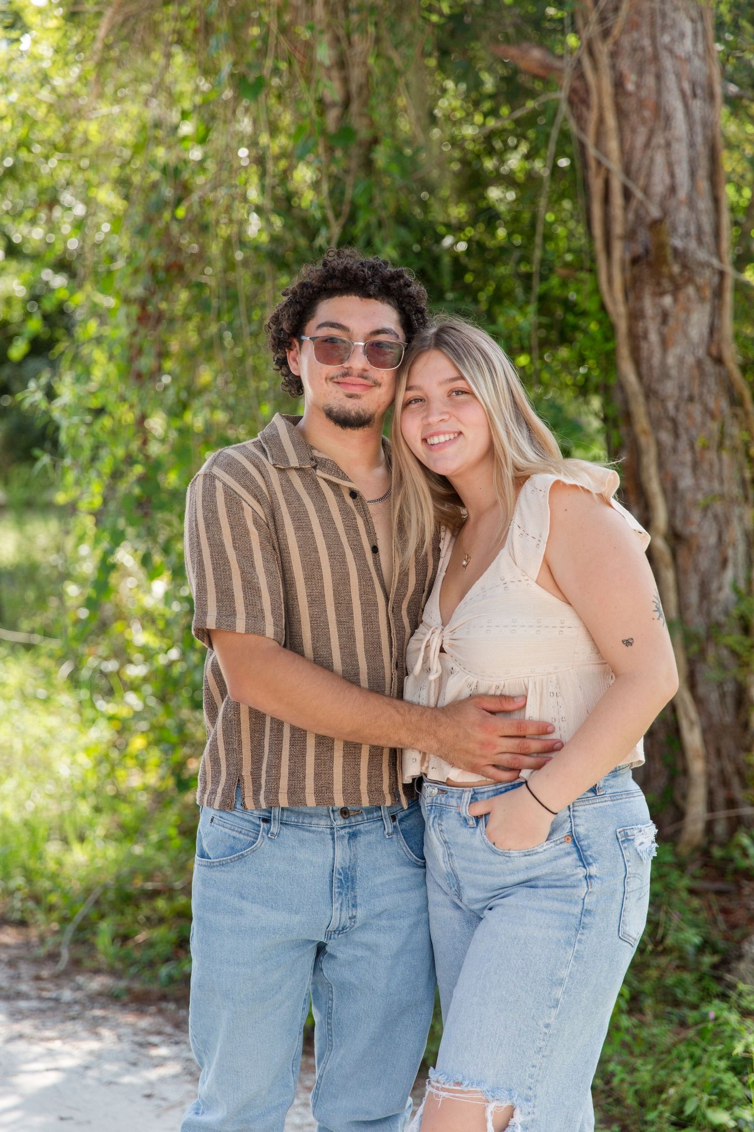 Young couple in casual jeans embracing and smiling outdoors surrounded by lush green trees.