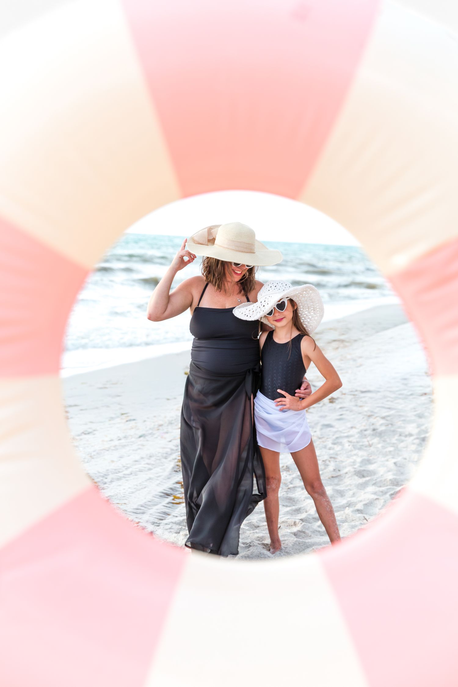 Mother and daughter in sun hats posing on beach seen through pink inflatable ring.