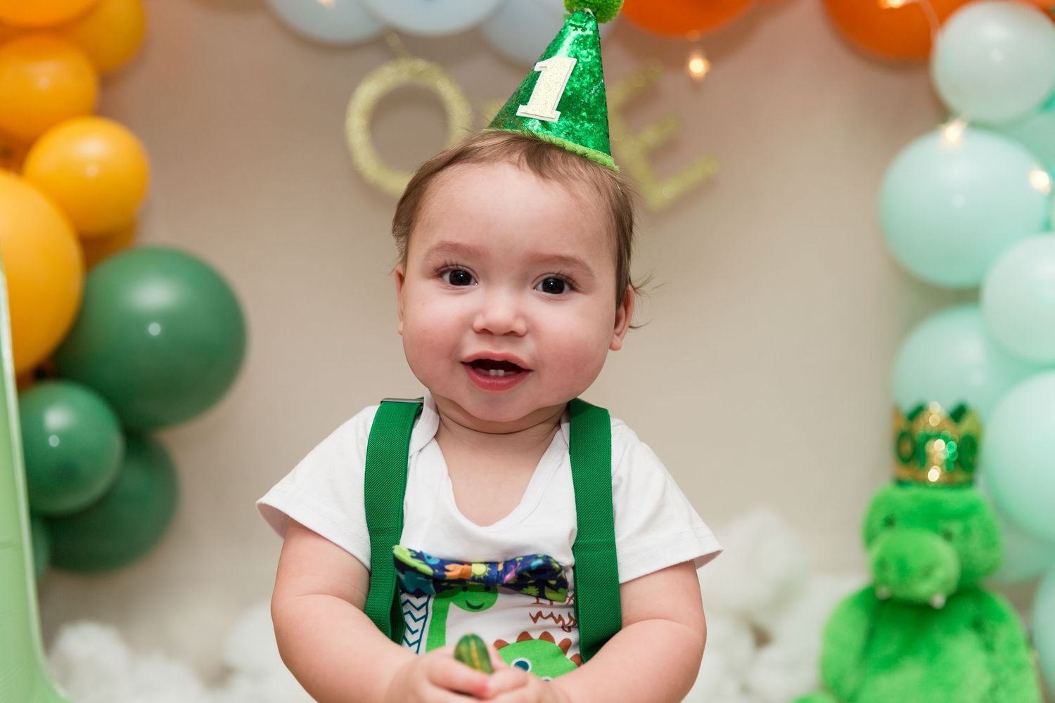 Smiling baby in green suspenders and birthday hat celebrates first birthday with colorful balloon decorations.
