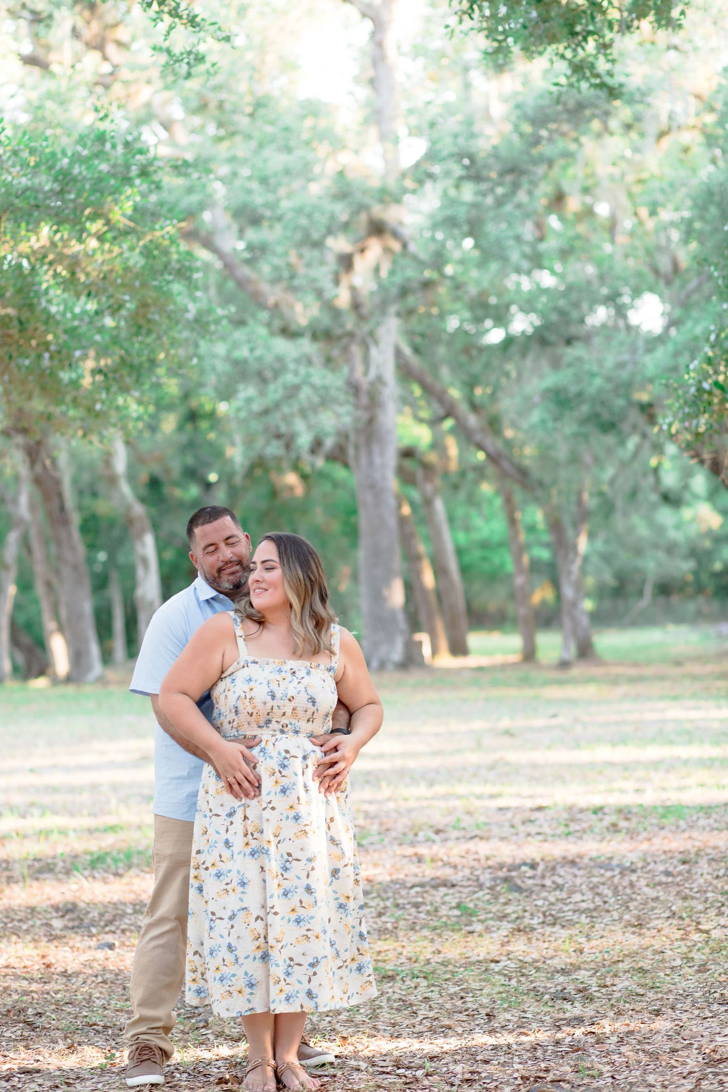 Couple embracing in a sunlit park, woman in floral midi dress, man in light blue shirt, surrounded by tall trees.
