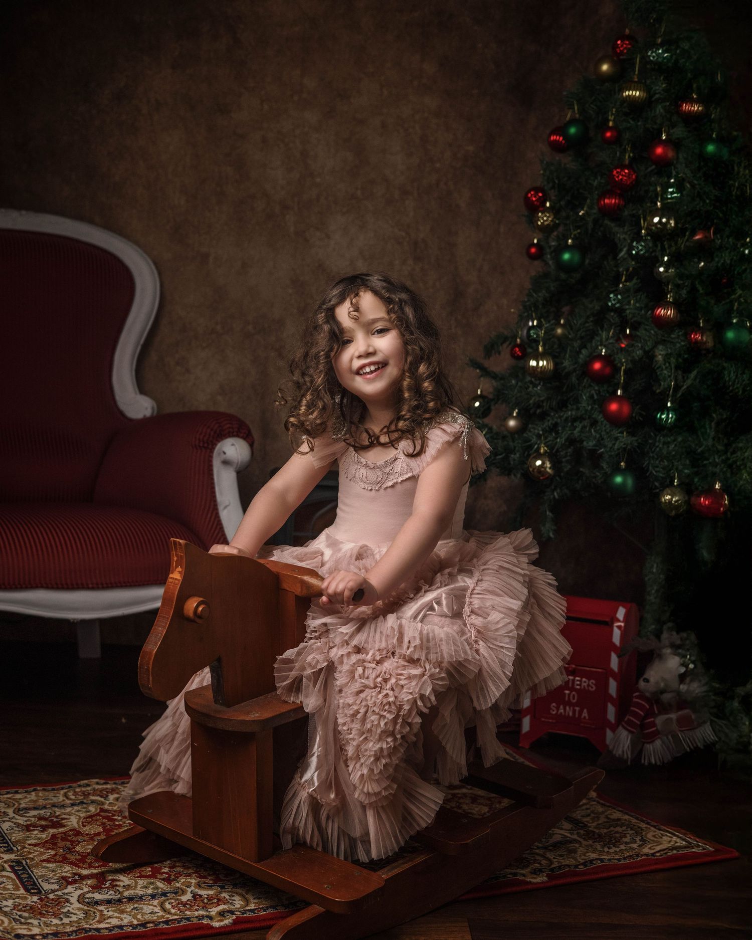 A child in a blush pink ruffle dress poses elegantly near a decorated Christmas tree in a cozy studio setting.