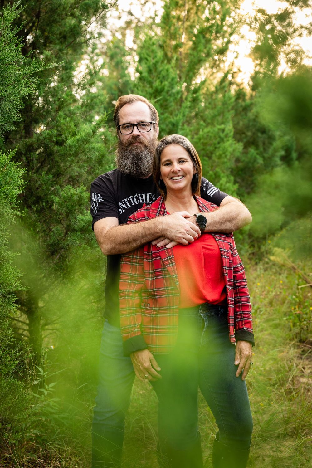 A couple embraces in a lush green outdoor setting wearing plaid and casual attire during a photo session.